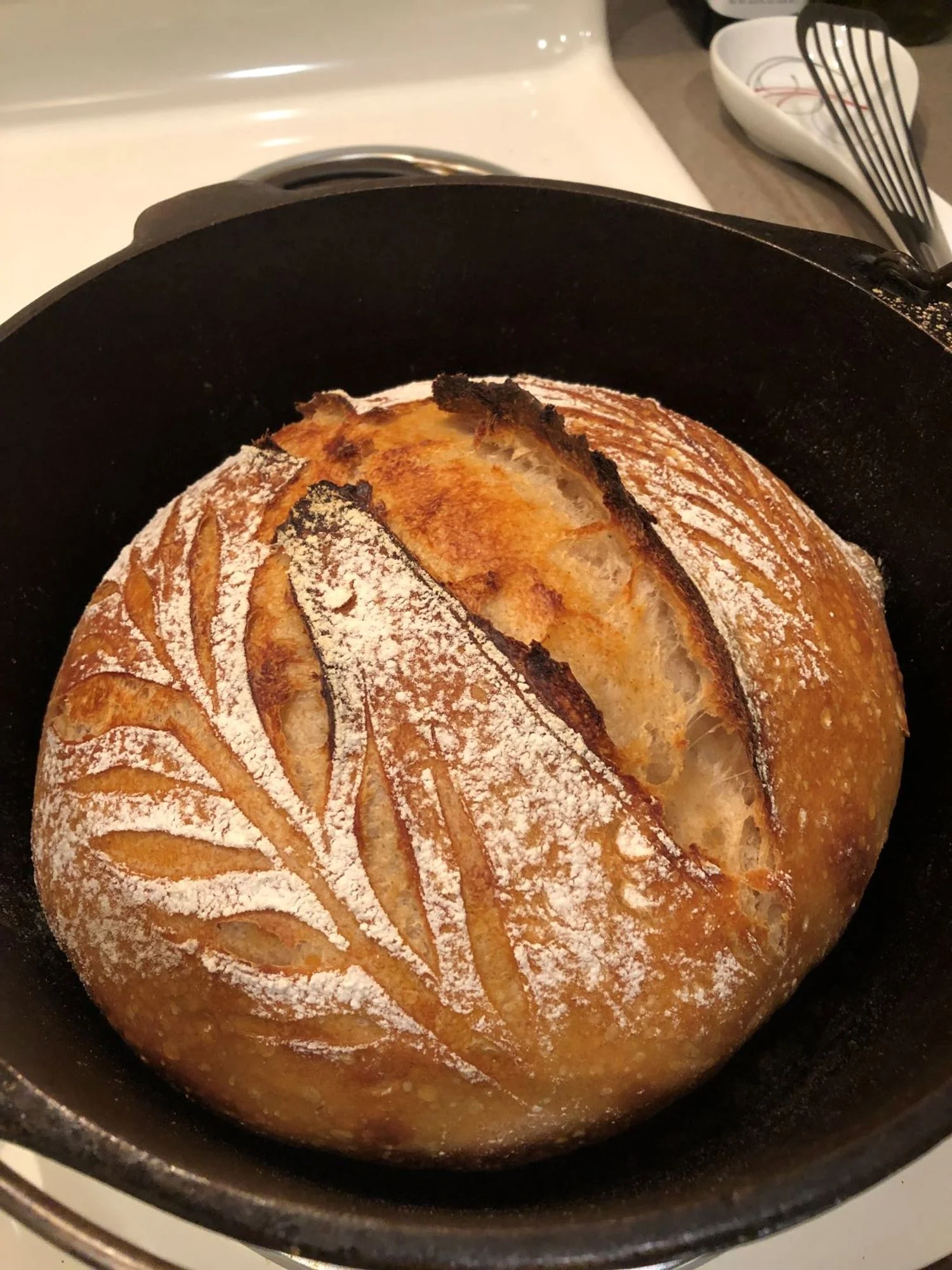Close-up of a round, freshly baked loaf of bread with a decorative scoring pattern on top, dusted with flour, inside a black cast iron skillet.