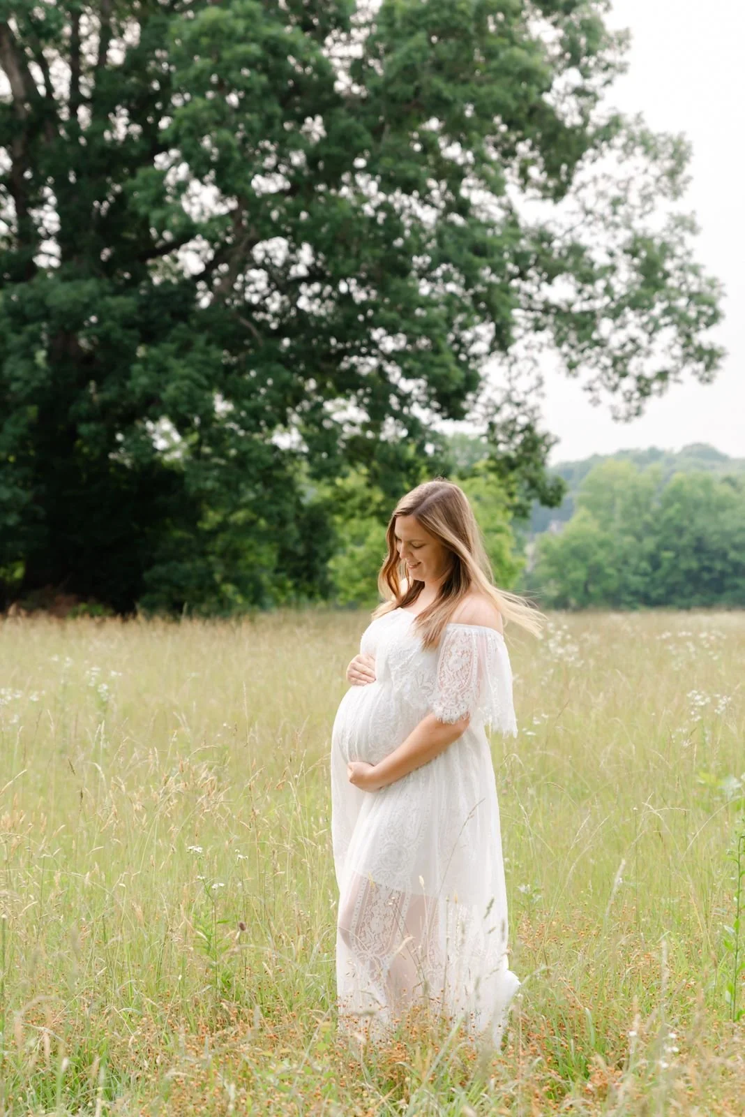 A pregnant woman in a white lace dress standing in a grassy field, holding her belly and smiling while looking down at it, with trees in the background.