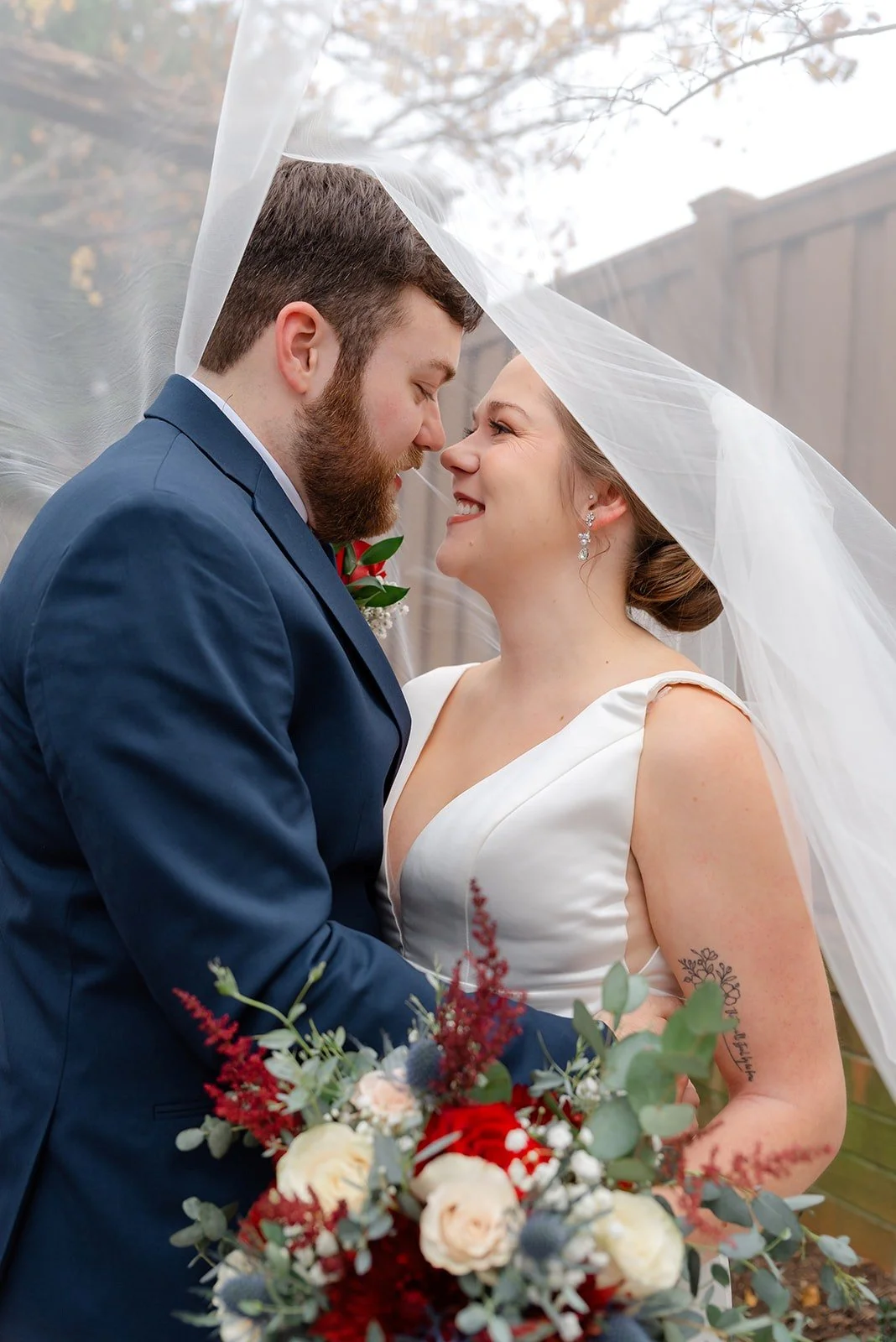 A bride and groom share a tender moment outdoors under a wedding veil. The groom wears a navy suit and the bride has a white dress, veil, and earrings. She holds a bouquet of red, white, and green flowers, and they smile intimately.
