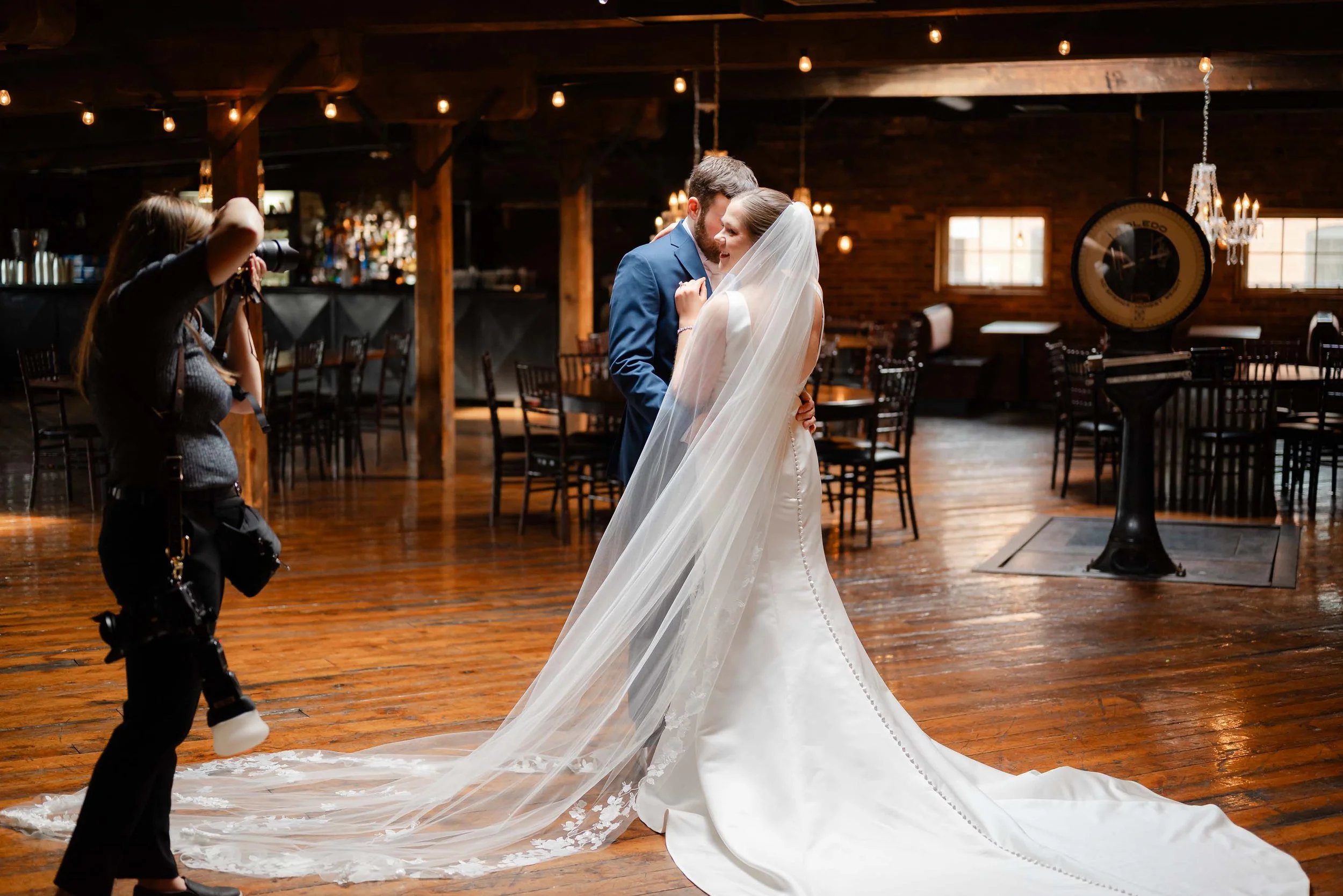 A bride and groom sharing a dance at their wedding reception, with a photographer capturing the moment inside a rustic venue with wooden floors and warm lighting.