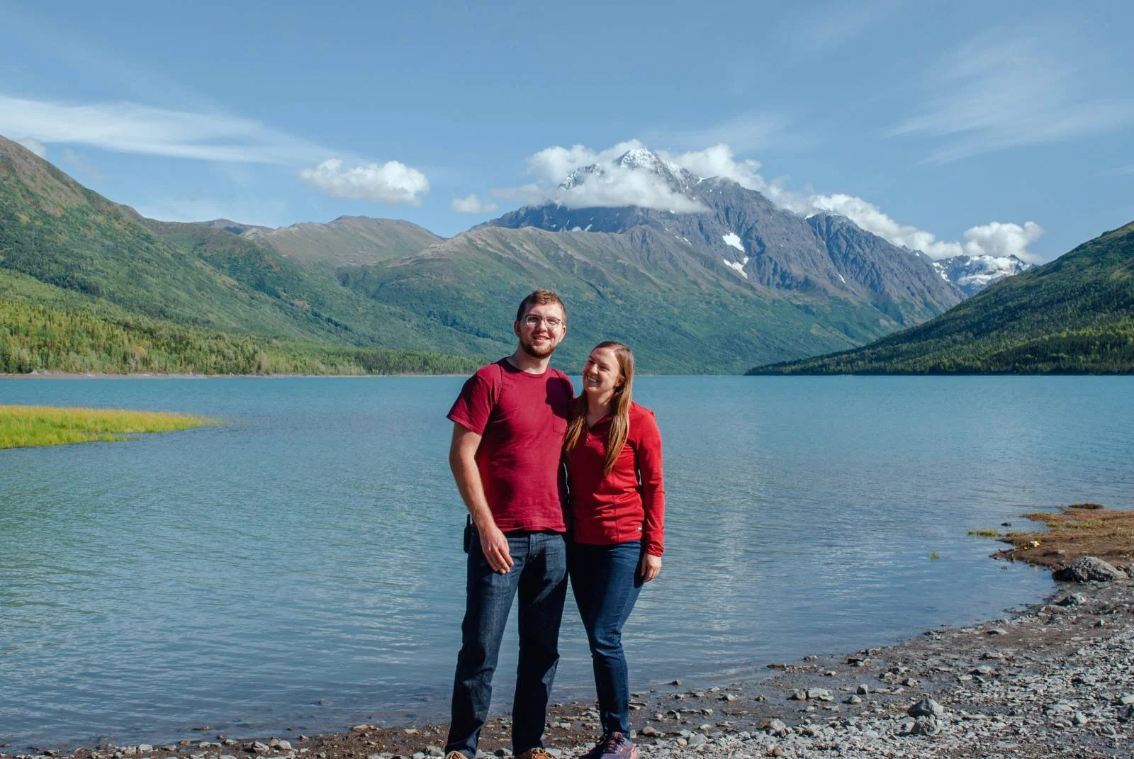 A couple standing on a rocky lakeshore, smiling, with a mountain range, a lake, and a partly cloudy sky in the background.