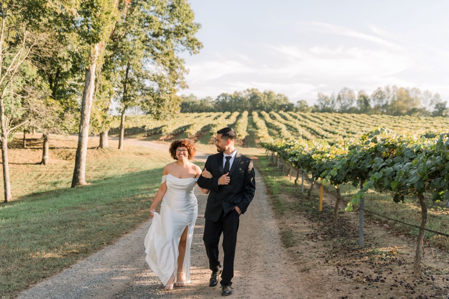 A couple in wedding attire walking on a dirt path through a vineyard with rows of grapevines, surrounded by trees, under a partly cloudy sky.