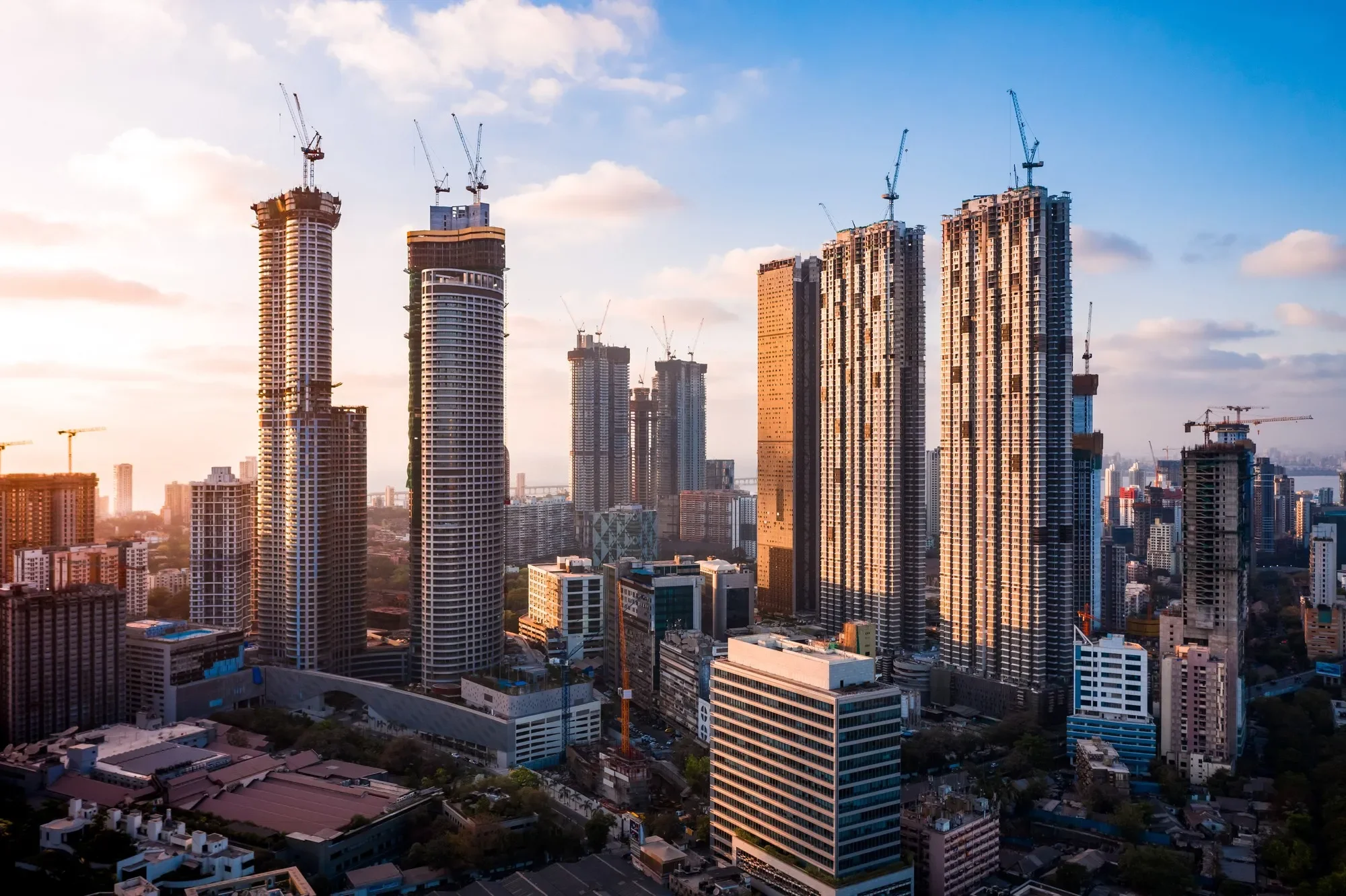 A city skyline during sunset showing many high-rise buildings, some under construction with cranes on top, and others completed. The sky is partly cloudy with warm light reflecting off the buildings.
