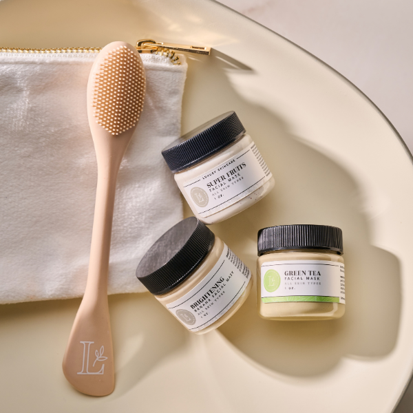 Skincare products and an exfoliating brush on a white tray, including a jar of super fruits facial mask, a jar of brightening mud, and a jar of green tea facial mask, with a white zippered pouch in the background.