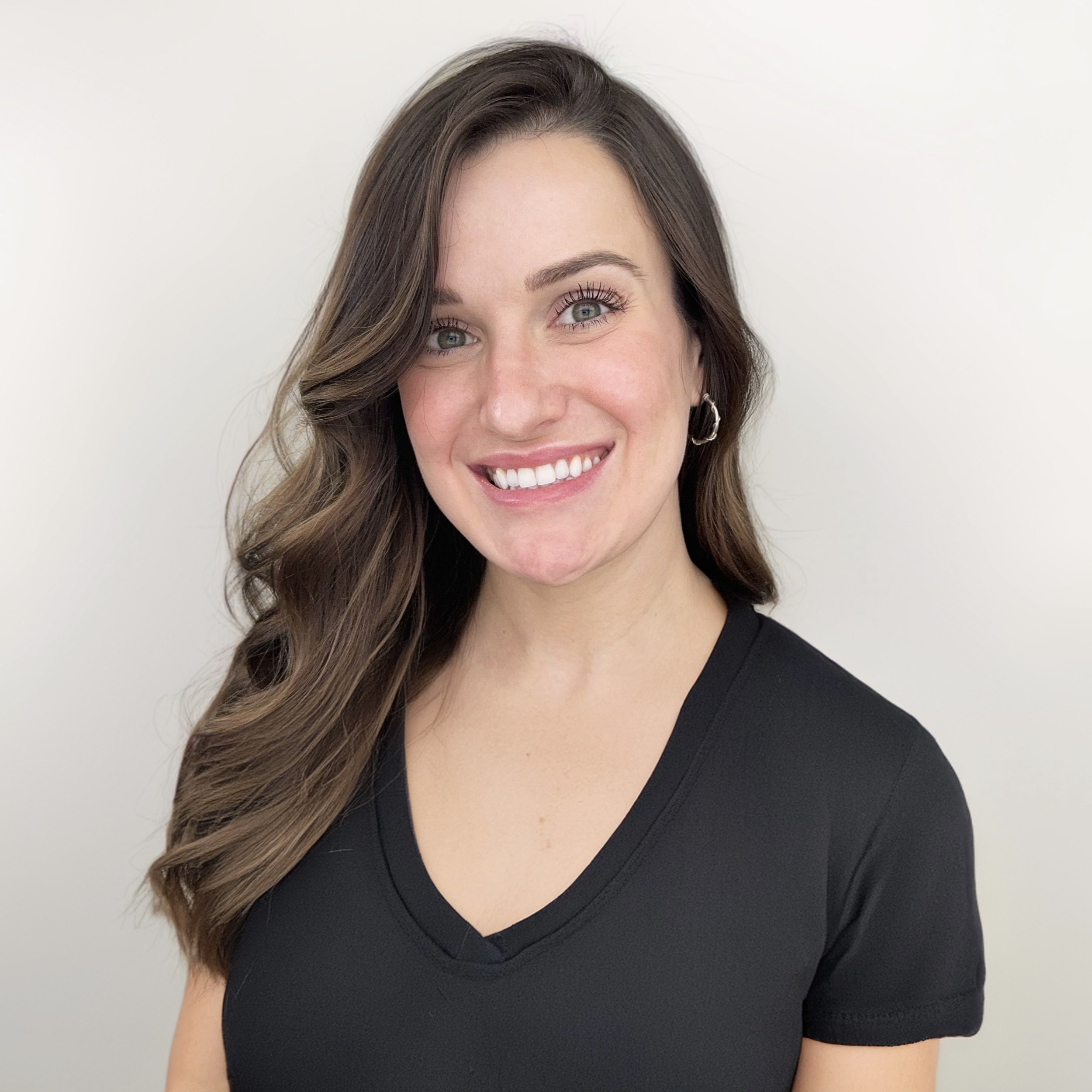 A young woman with long wavy brown hair, blue eyes, and a bright smile wearing a black V-neck shirt and small silver hoop earrings, standing against a plain light-colored background.