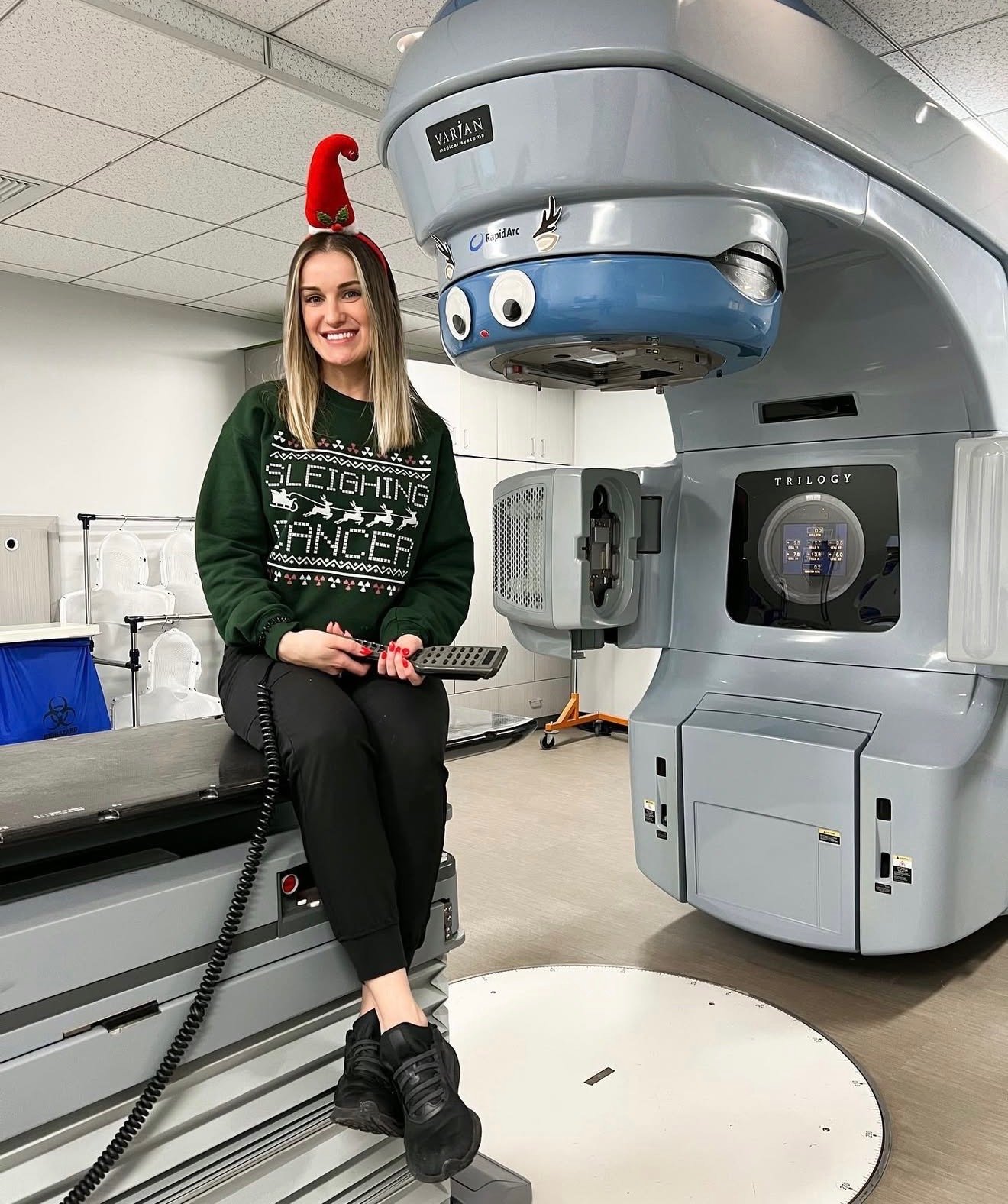 A woman sitting on a medical imaging table in a hospital, wearing a green sweater with Christmas designs and the words 'Sleighing Cancer,' a red Christmas hat with a pom-pom, and holding a remote. She is smiling, and a large medical scanner is positioned next to her.