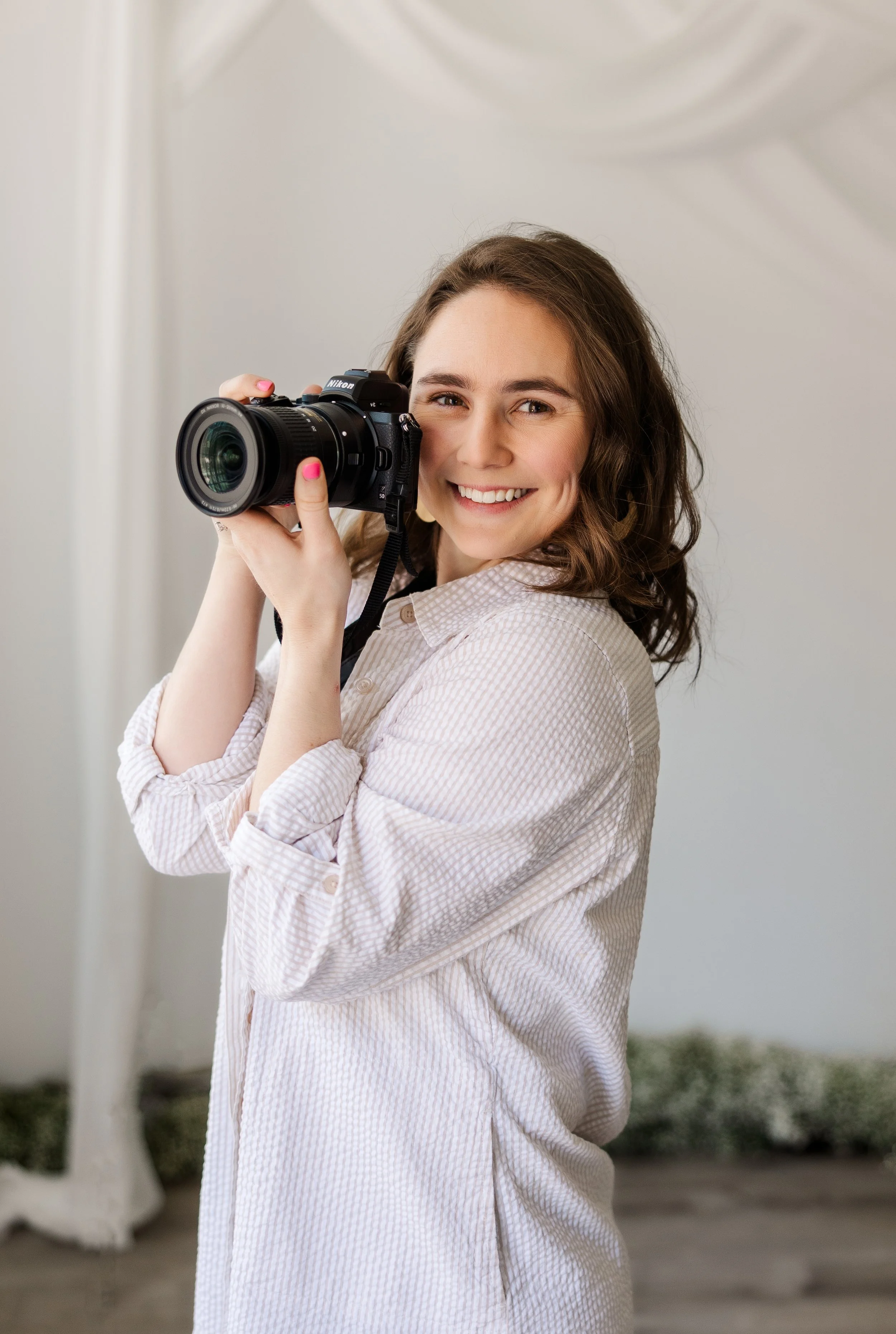 A young woman with wavy brown hair smiling and holding a camera, wearing a light-colored, checkered shirt in a bright indoor setting.