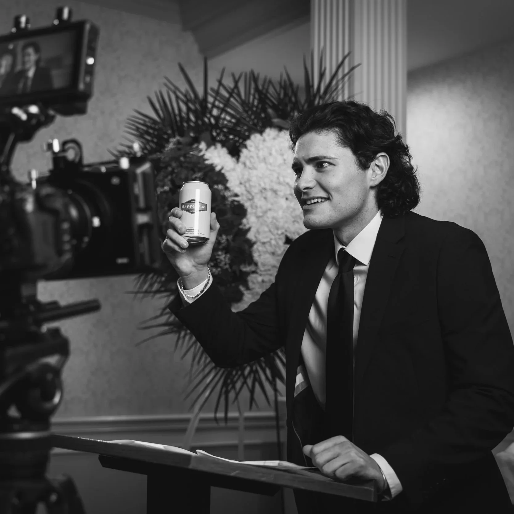 Black and white behind the scenes photo of actor Maxwell Morris in a suit and tie holding a Happy Dad can during the shooting of A Happy Dad Funeral, with a camera and floral arrangement in the background.