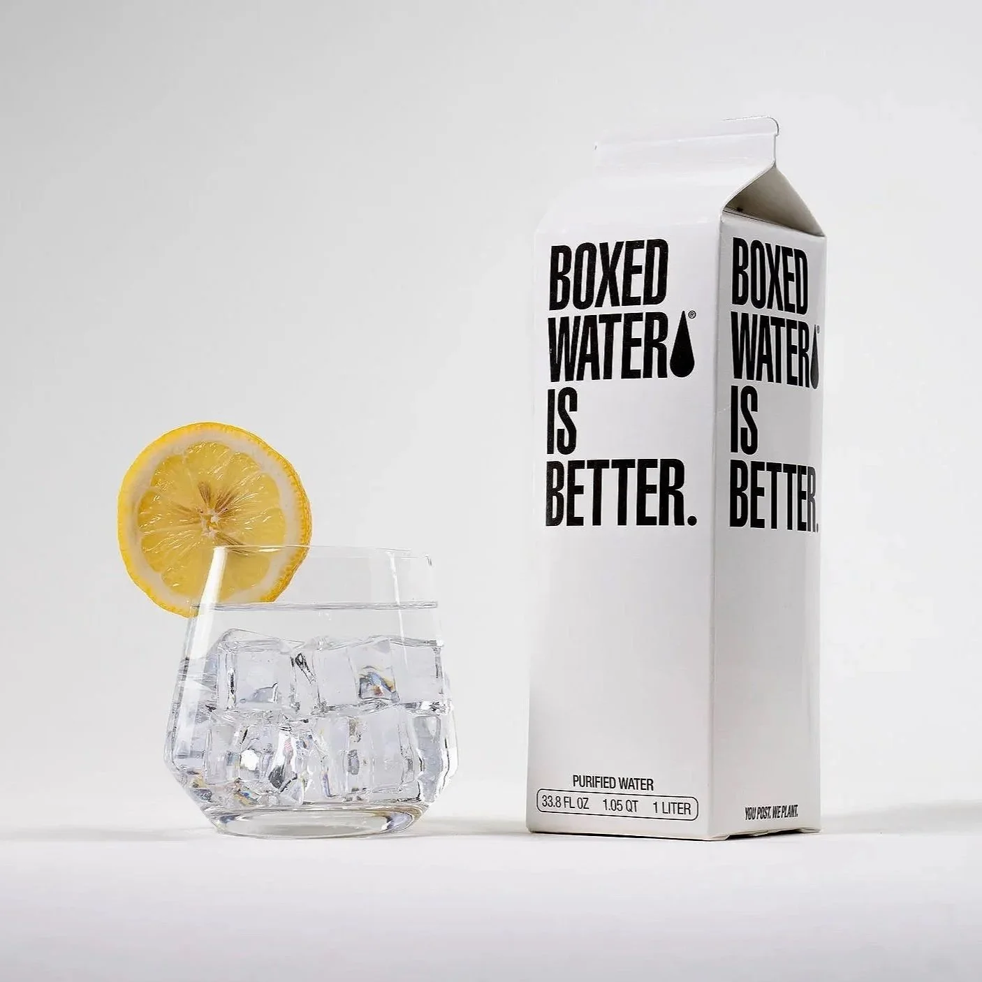 A glass of water with ice and a lemon slice on the rim next to a carton of boxed water with the text "Boxed Water is Better." on a white background.