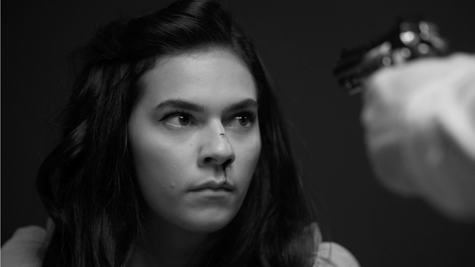 A woman with dark, wavy hair looking intently at a makeup artist applying eyeliner with a brush.