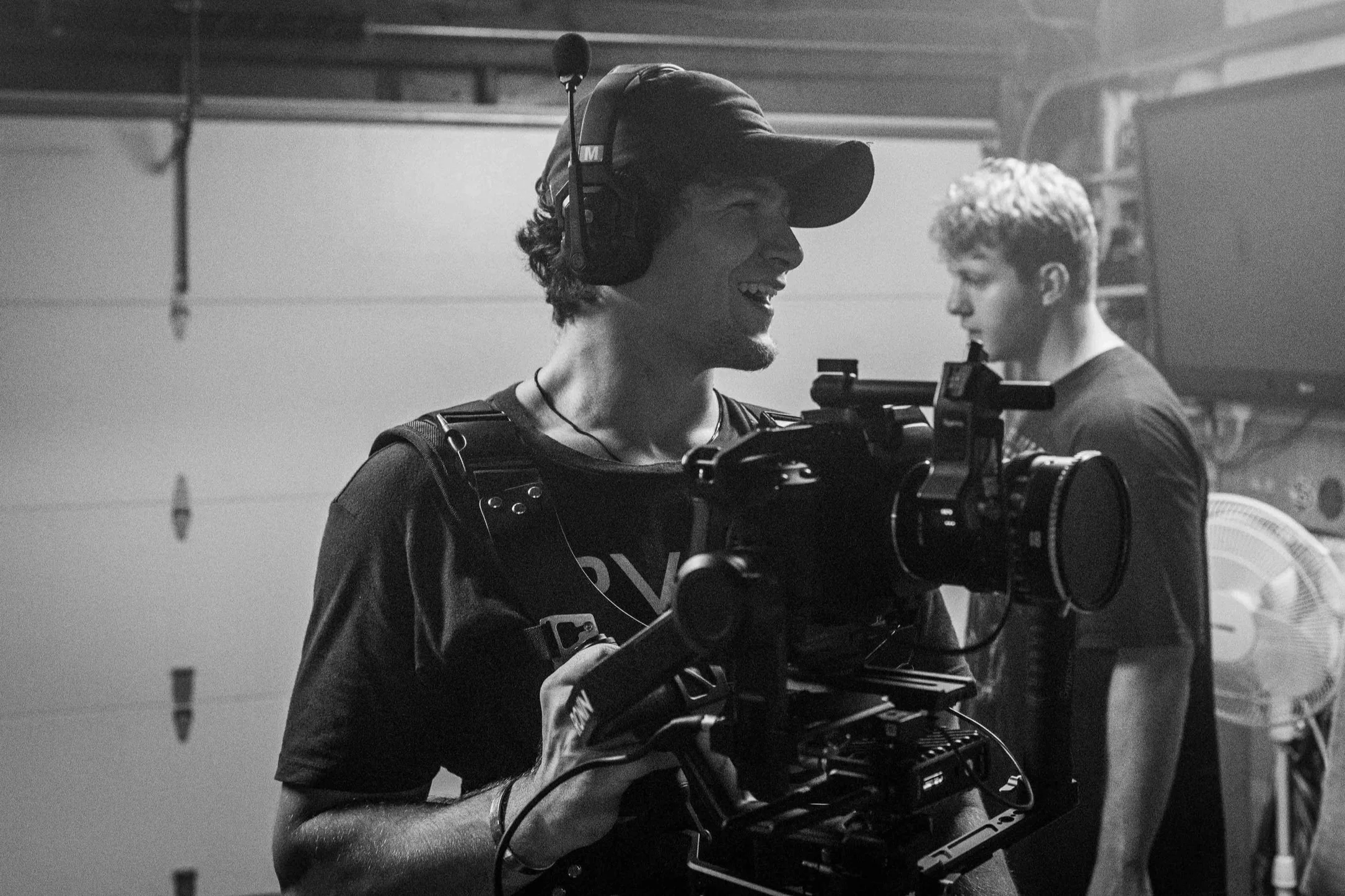 Black-and-white photo of a young man with curly hair, wearing a cap and headphones, operating a professional camera rig in a garage or workshop setting. Another young man is in the background, looking at something.