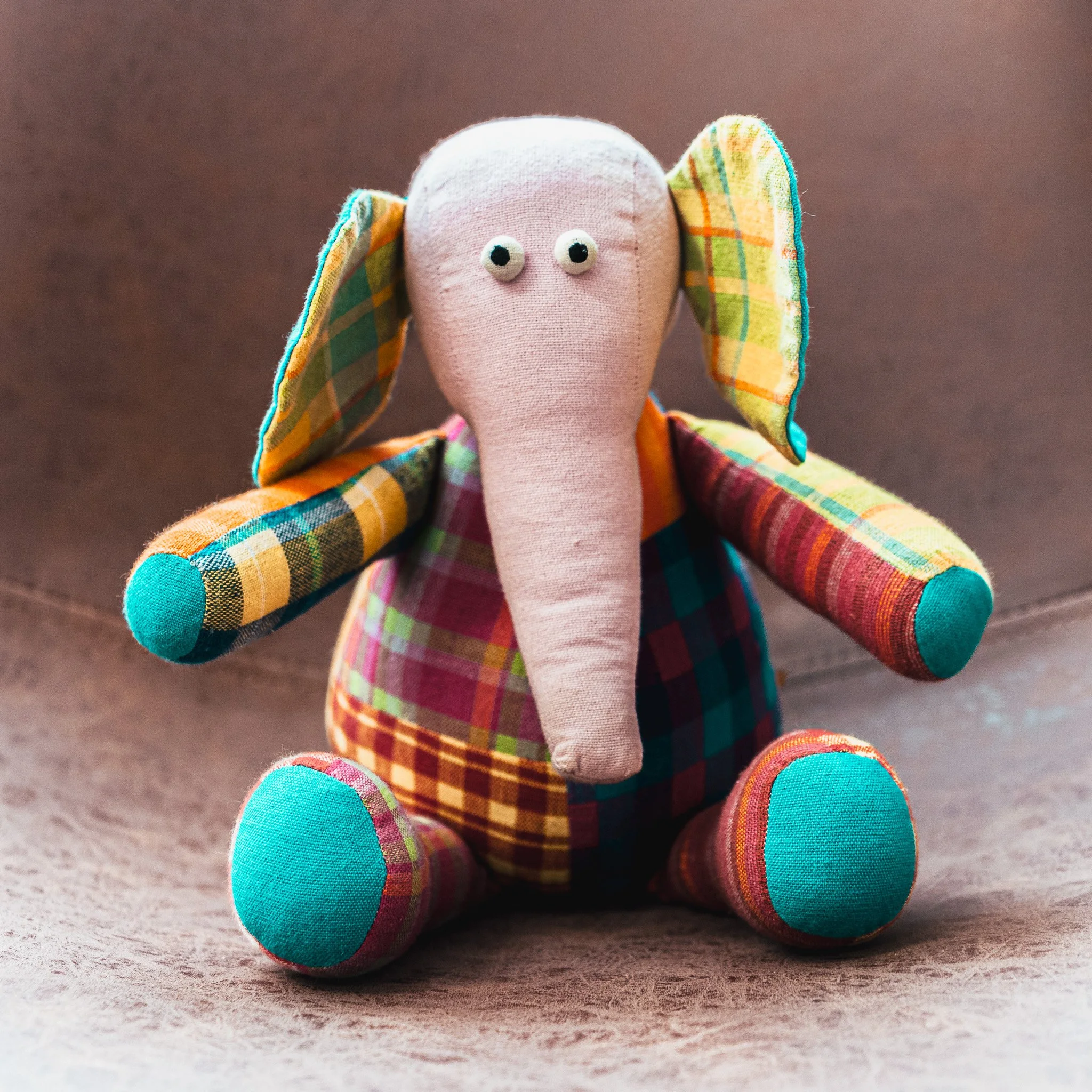 Colorful fabric stuffed toy elephant with large floppy ears, small eyes, and a long trunk, sitting on a beige surface.
