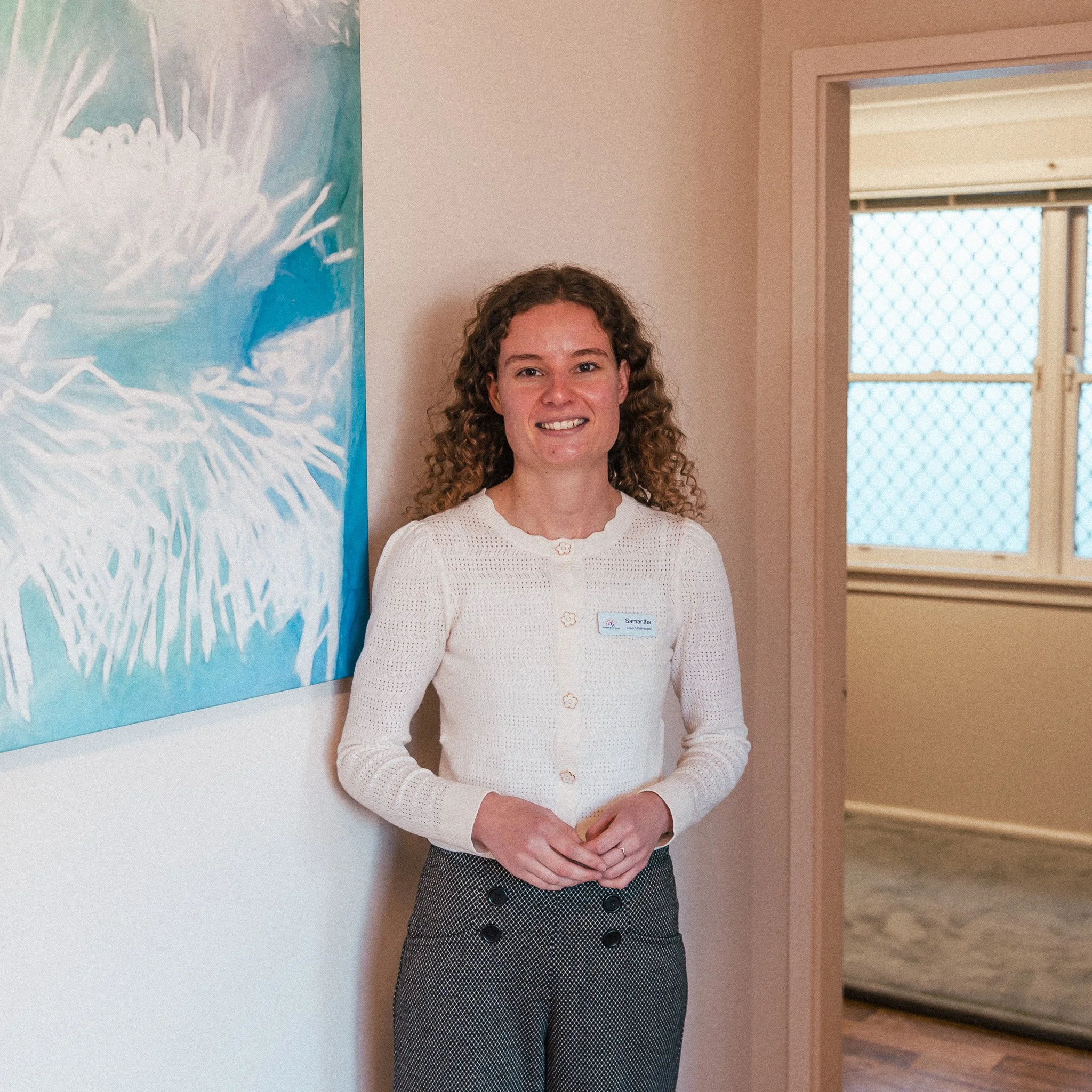 A woman with curly brown hair standing next to a colorful painting on the wall, wearing a white cardigan and gray patterned pants, smiling at the camera.
