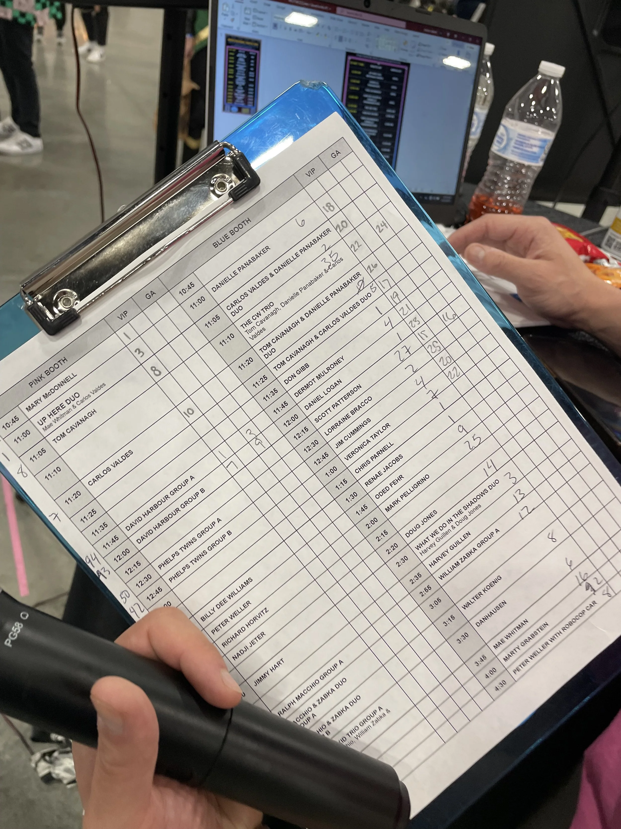 A person holding a clipboard with a schedule or event list at an indoor event or convention, with a laptop, water bottle, and snacks visible in the background.