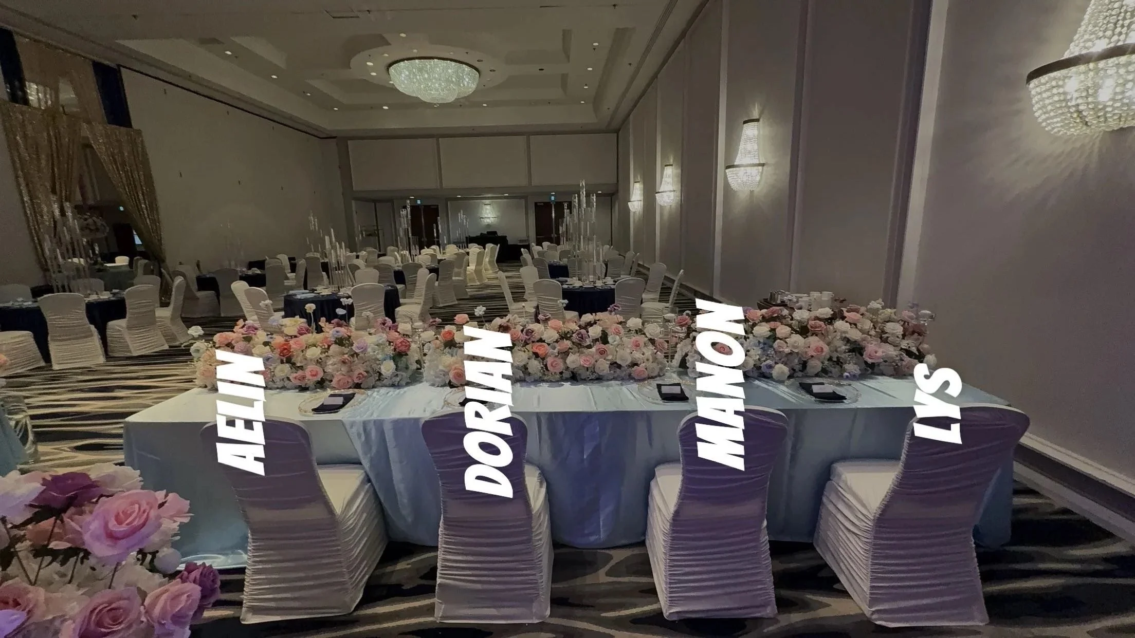 Elegant banquet hall with a long table decorated with pink and white flower arrangements, surrounded by white chairs with pleated covers, and a high ceiling with a large chandelier.