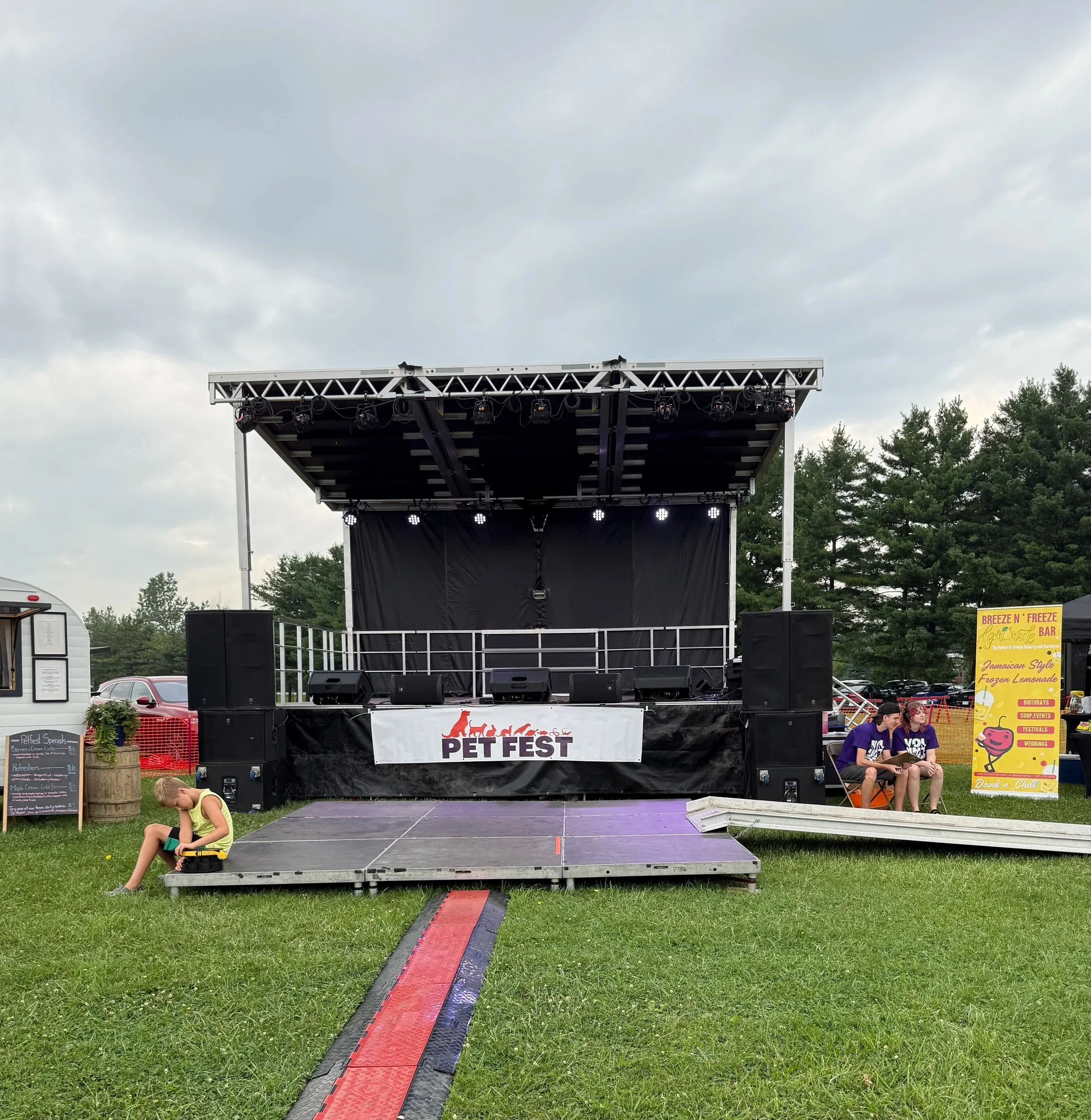 An outdoor pet festival stage with a sign that reads 'Pet Fest' and a purple carpet extending towards it, surrounded by a grassy area, trees, and cloudy skies, with two children sitting on chairs and a girl in a yellow shirt sitting on the grass.