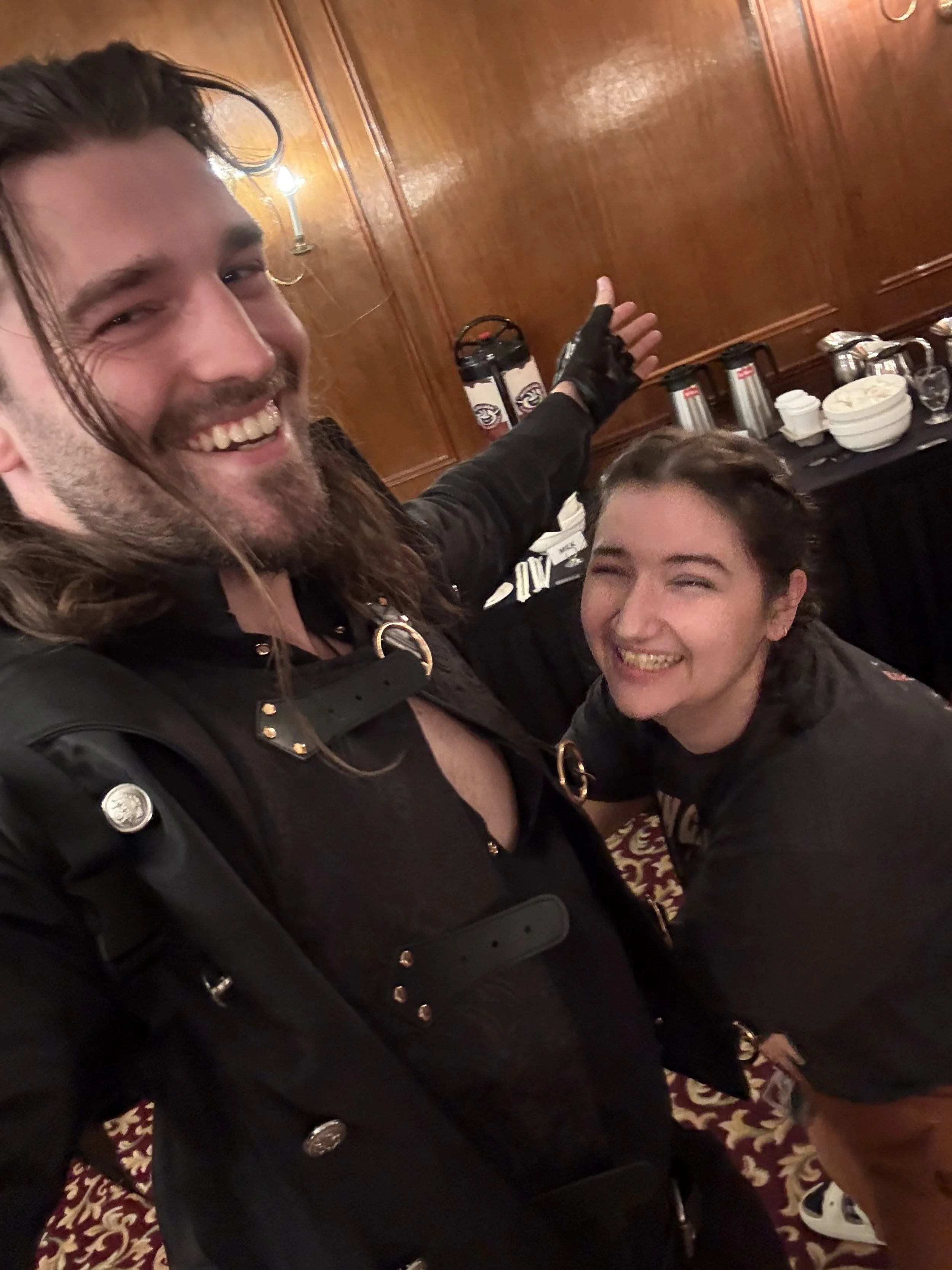 Two people smiling and posing for a selfie in a room with wooden-paneled walls. The person on the left has long brown hair and a beard, wearing a black leather outfit with belt accents. The person on the right has short, dark, curly hair, wearing a d