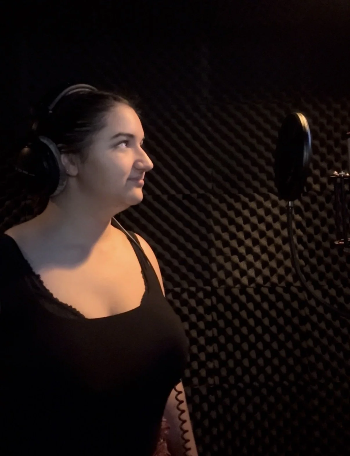 A woman with dark hair pulled back, wearing headphones, standing in a recording studio with black acoustic foam panels, looking at a microphone with a pop filter.