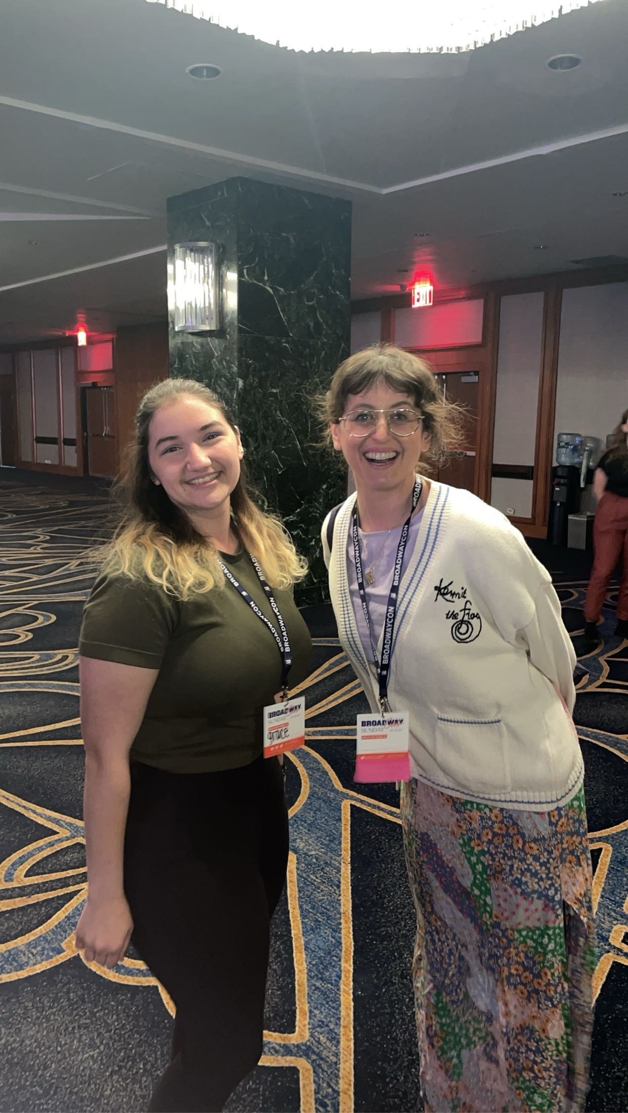 Two women smiling in a hotel lobby with a patterned carpet, dark marble pillar, and wood-paneled walls. They are wearing conference badges.