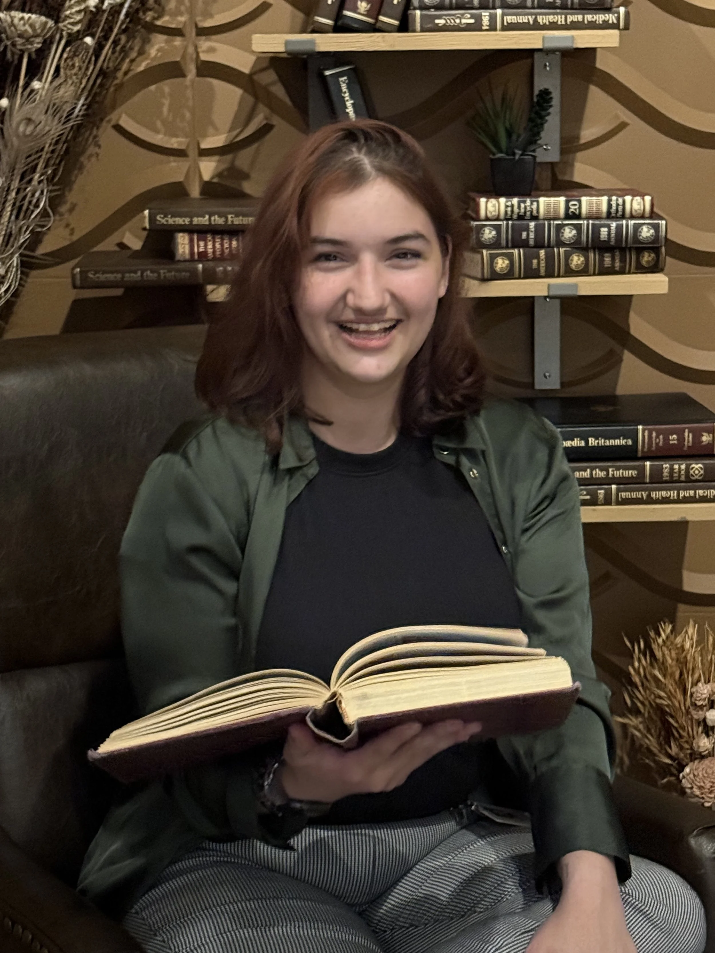 A woman with shoulder-length brown hair, wearing a black shirt and green jacket, sitting on a brown chair and holding an open book, smiling in a library or bookstore setting with shelves of books behind her.