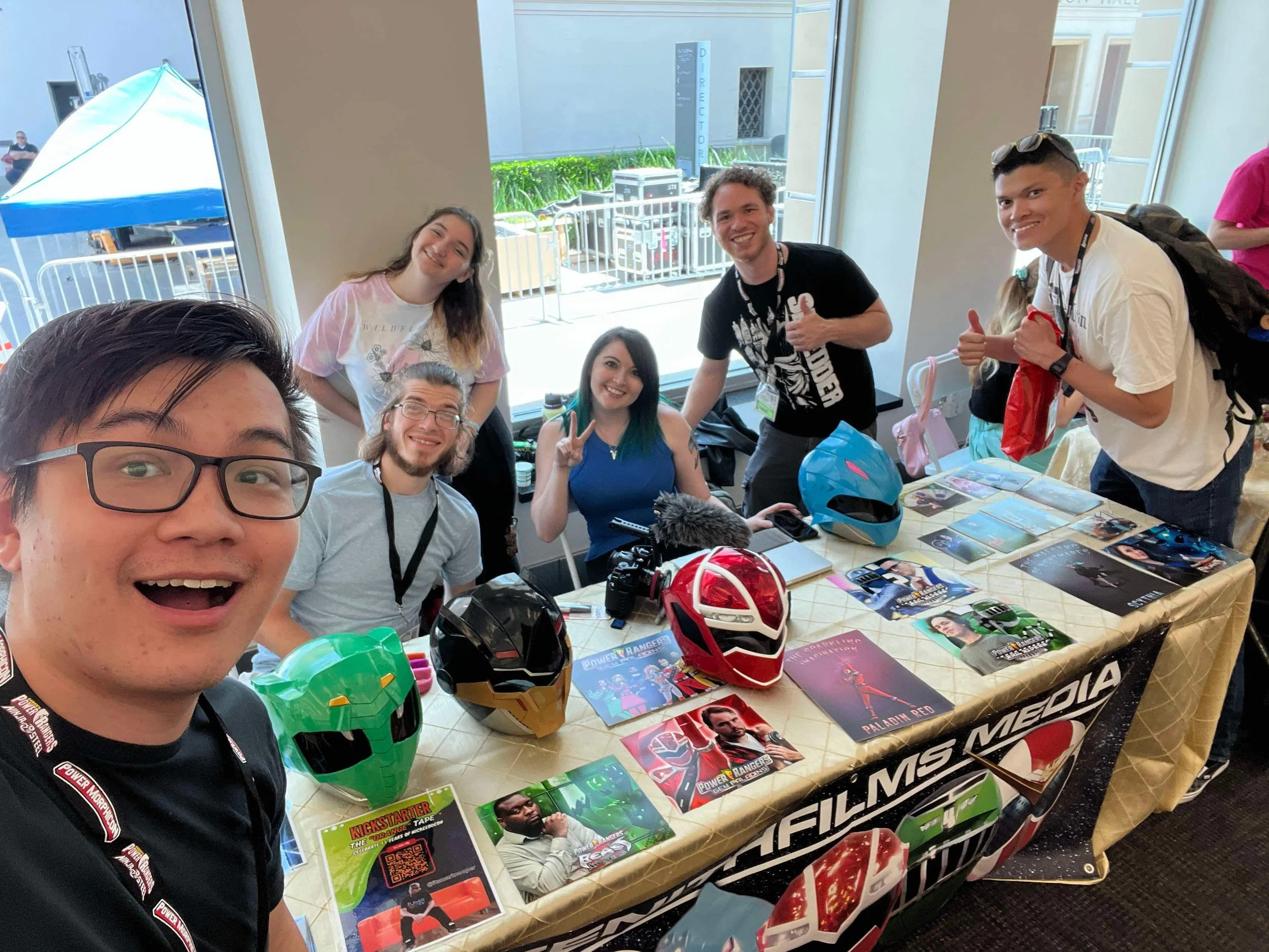 Group of people at a promotional table with Power Rangers helmets and posters, smiling and posing for a photo.