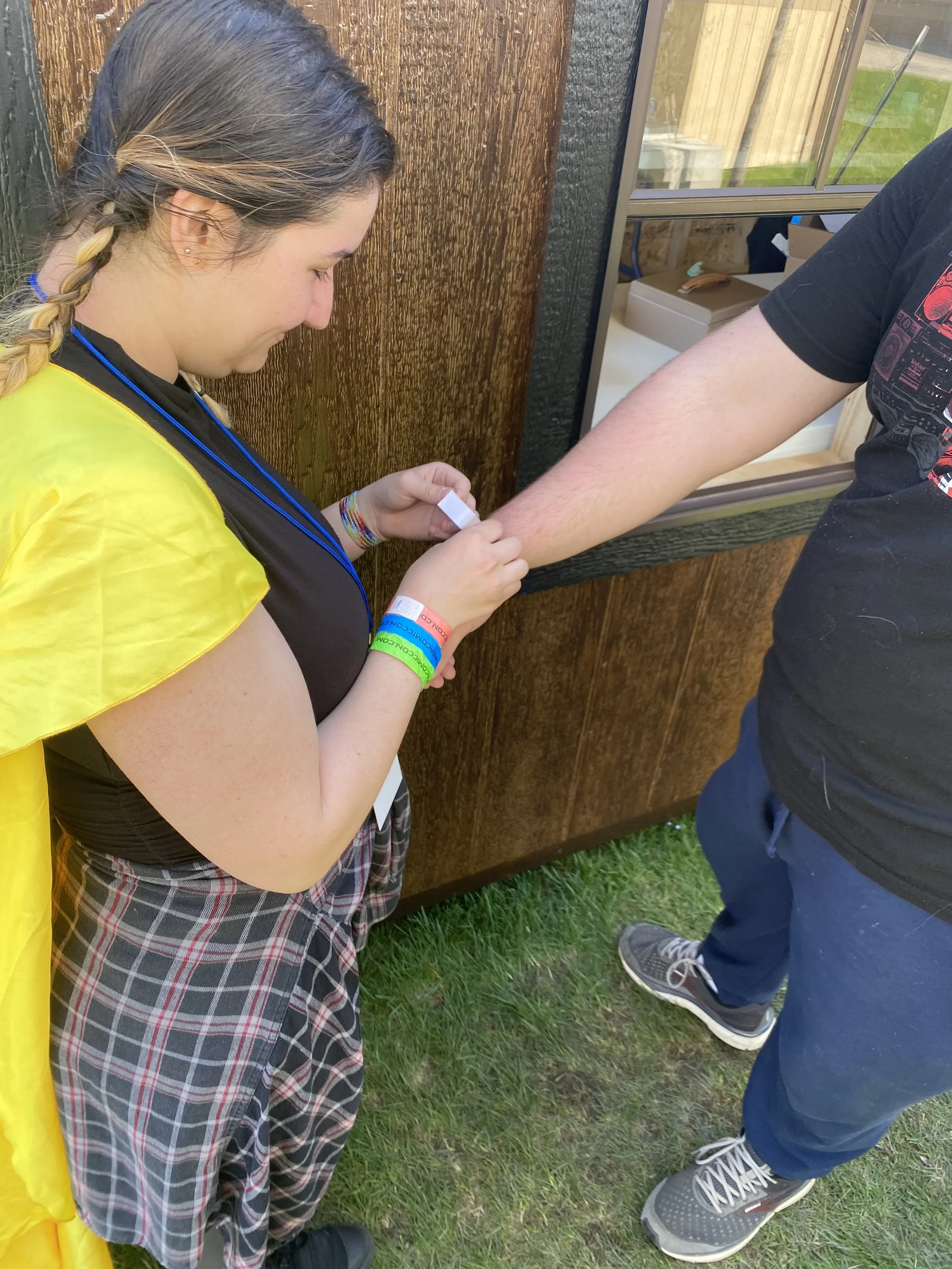 A young girl in a yellow cape and plaid skirt is tattooing a person's arm outdoors near a wooden fence and grass. She has a braid in her hair and colorful bracelets on her wrist.