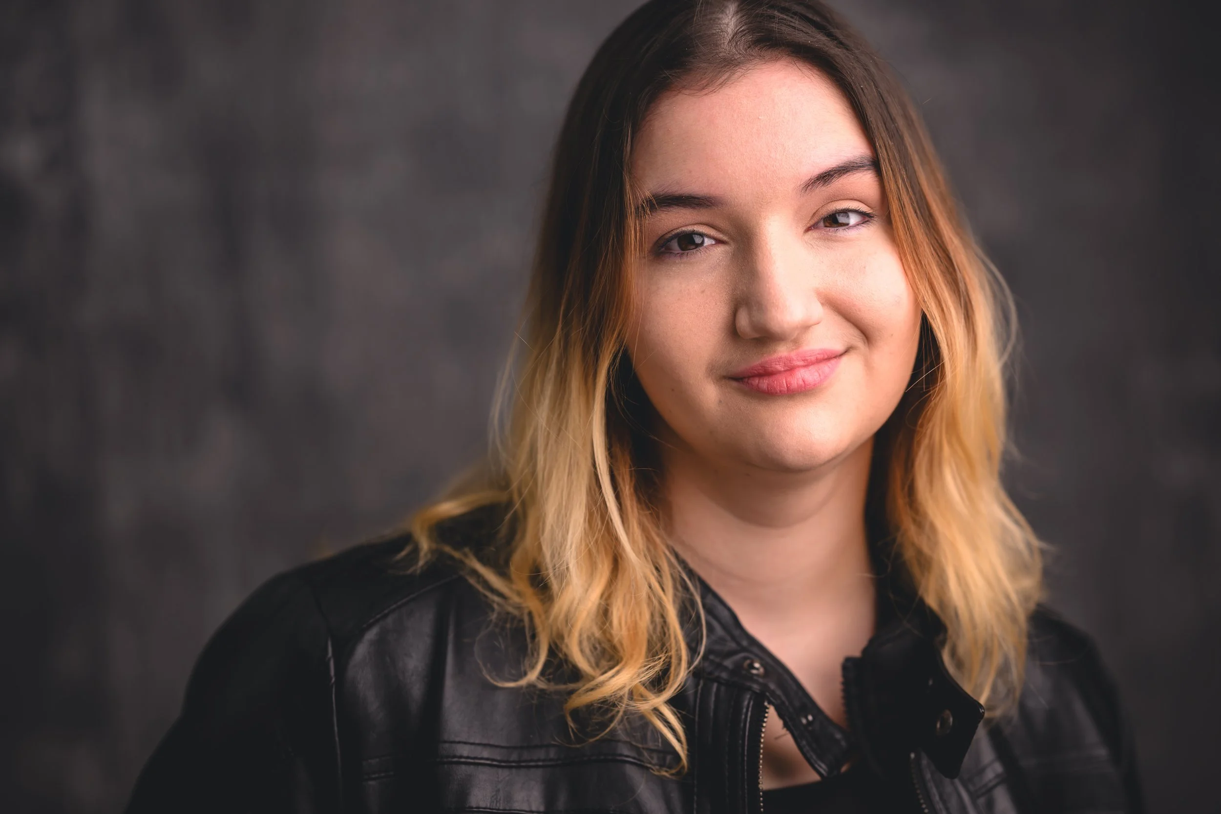 A young woman with blonde and brown hair smiling, wearing a black leather jacket, against a dark blurred background.
