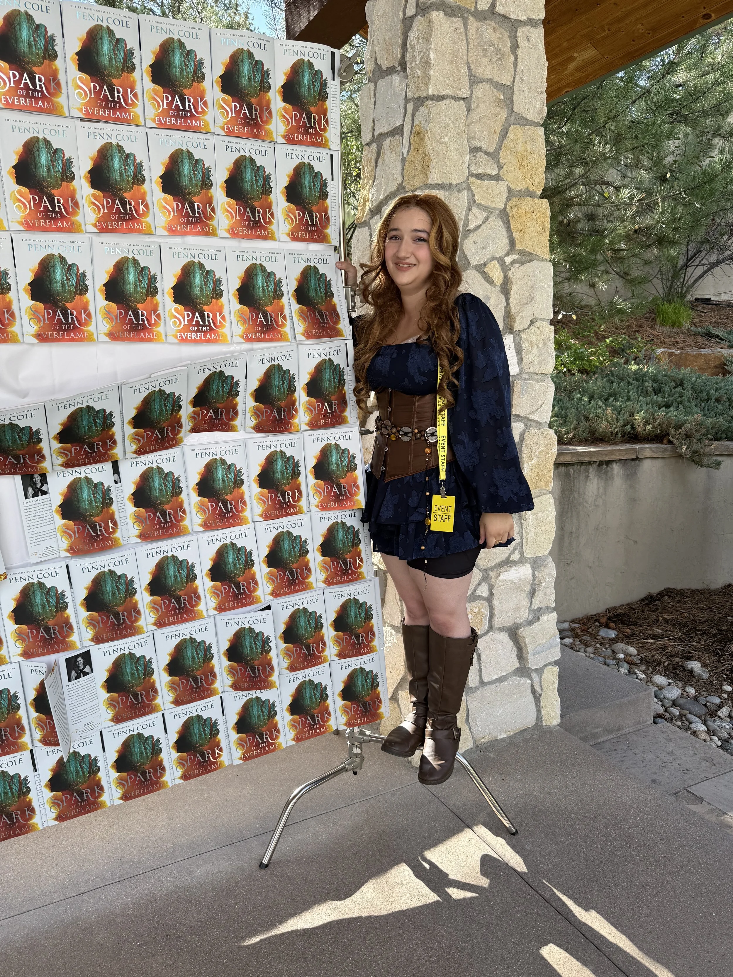 Young woman dressed as a fantasy character standing on a small platform, smiling, in front of a wall covered with copies of the book "Spark of the Everflame" by Penn Cole, outdoors with trees and stone wall in the background.
