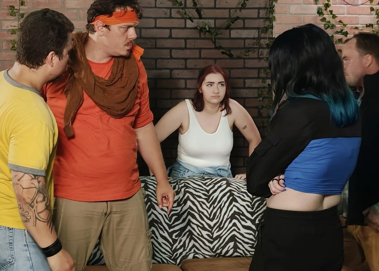 Five people are arguing in a room with a brick wall and some green vines. One woman with red hair and a white tank top appears upset and is standing behind a zebra print table. The other four people are men and women standing closely, angry, and engaged in a heated discussion.