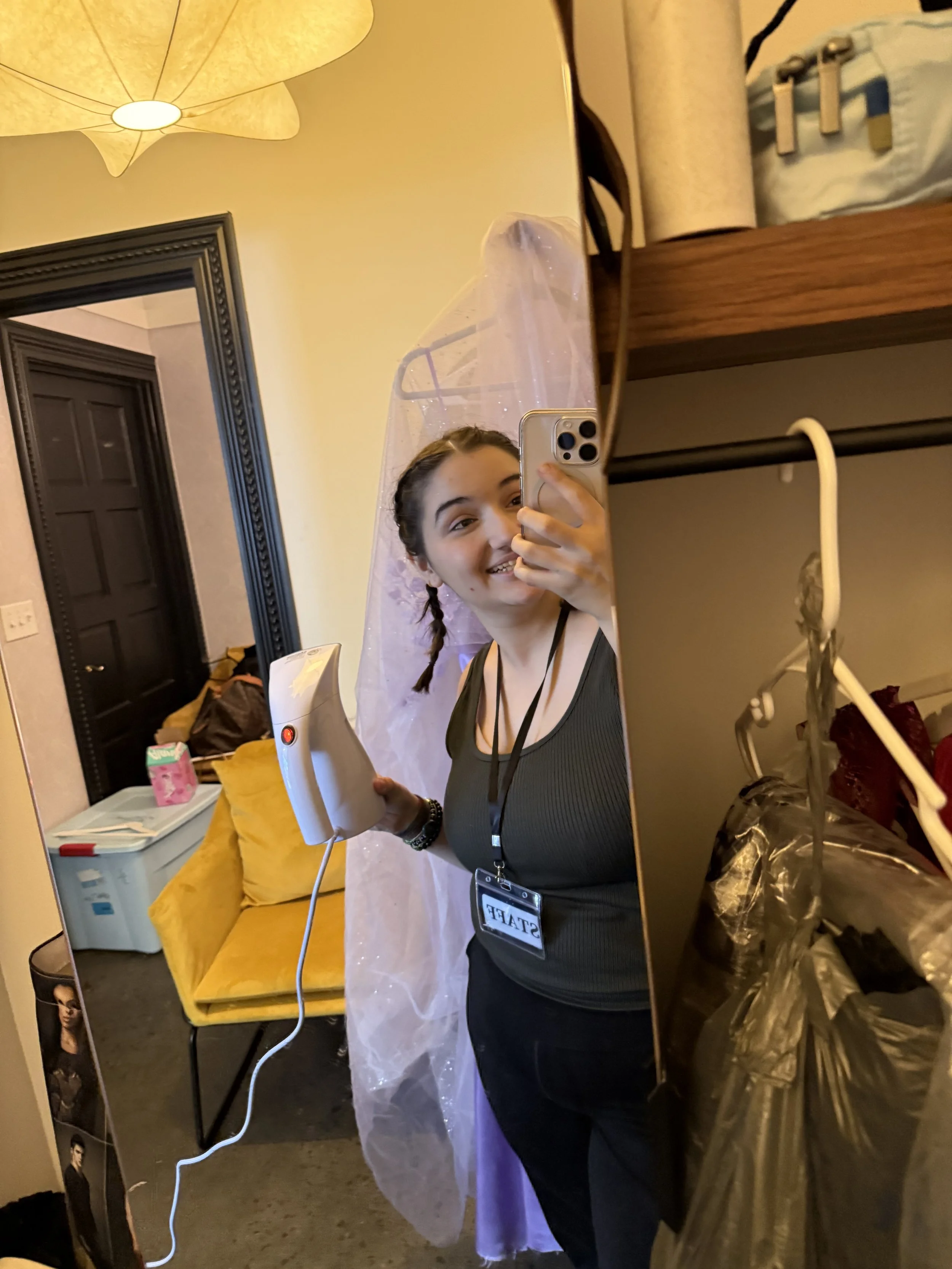 A woman taking a mirror selfie in a room, smiling, holding a handheld steamer or clothes steamer, wearing a black top and a lanyard with a badge that reads 'STAFF'. There is a yellow chair with a yellow pillow, a door frame, and various items on shel