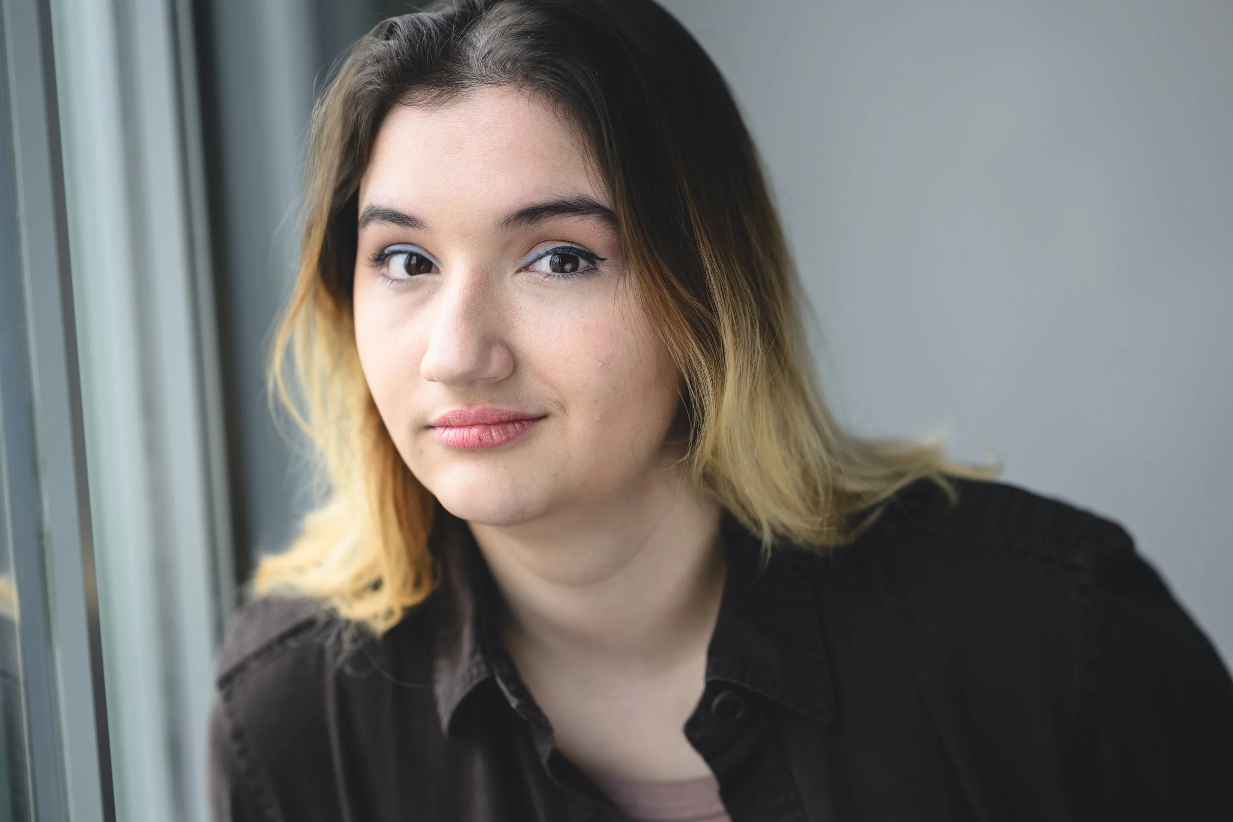 A young woman with light olive skin and shoulder-length ombre hair, wearing a brown shirt, sitting near a window with a blurred background. Professional Headshot by Matt Simpkins, 2024