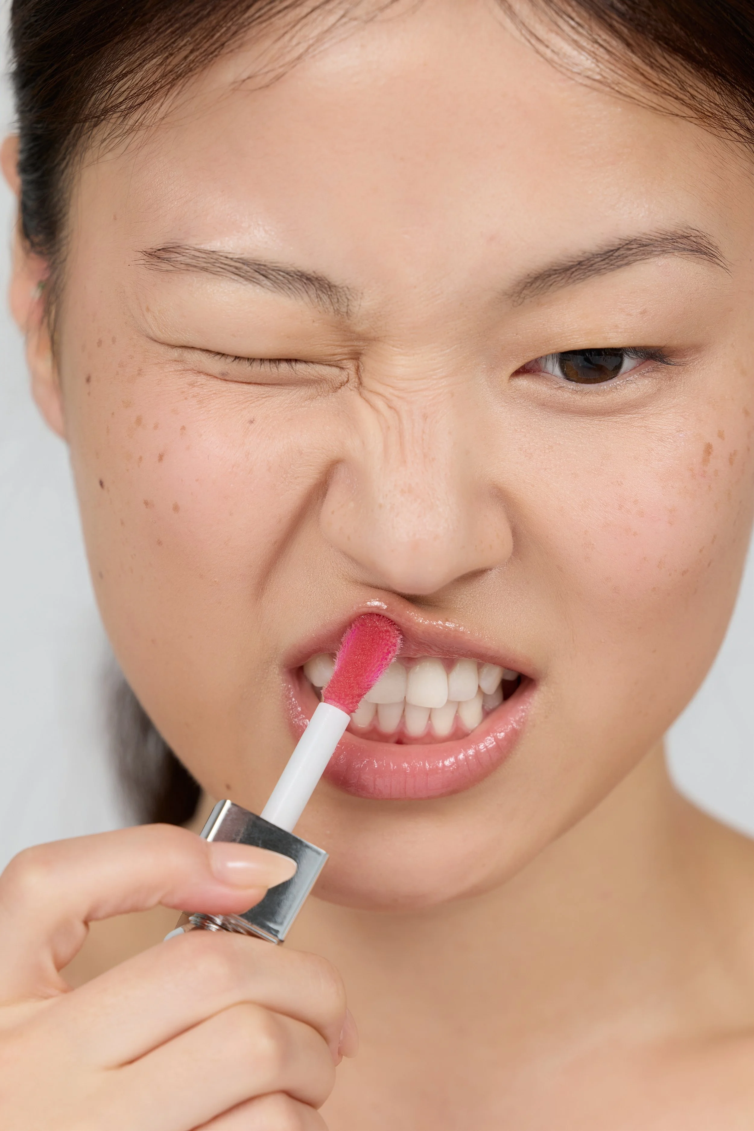 A woman with light skin and brown hair applying pink lip gloss, smiling with her teeth showing, while holding a lip gloss tube.