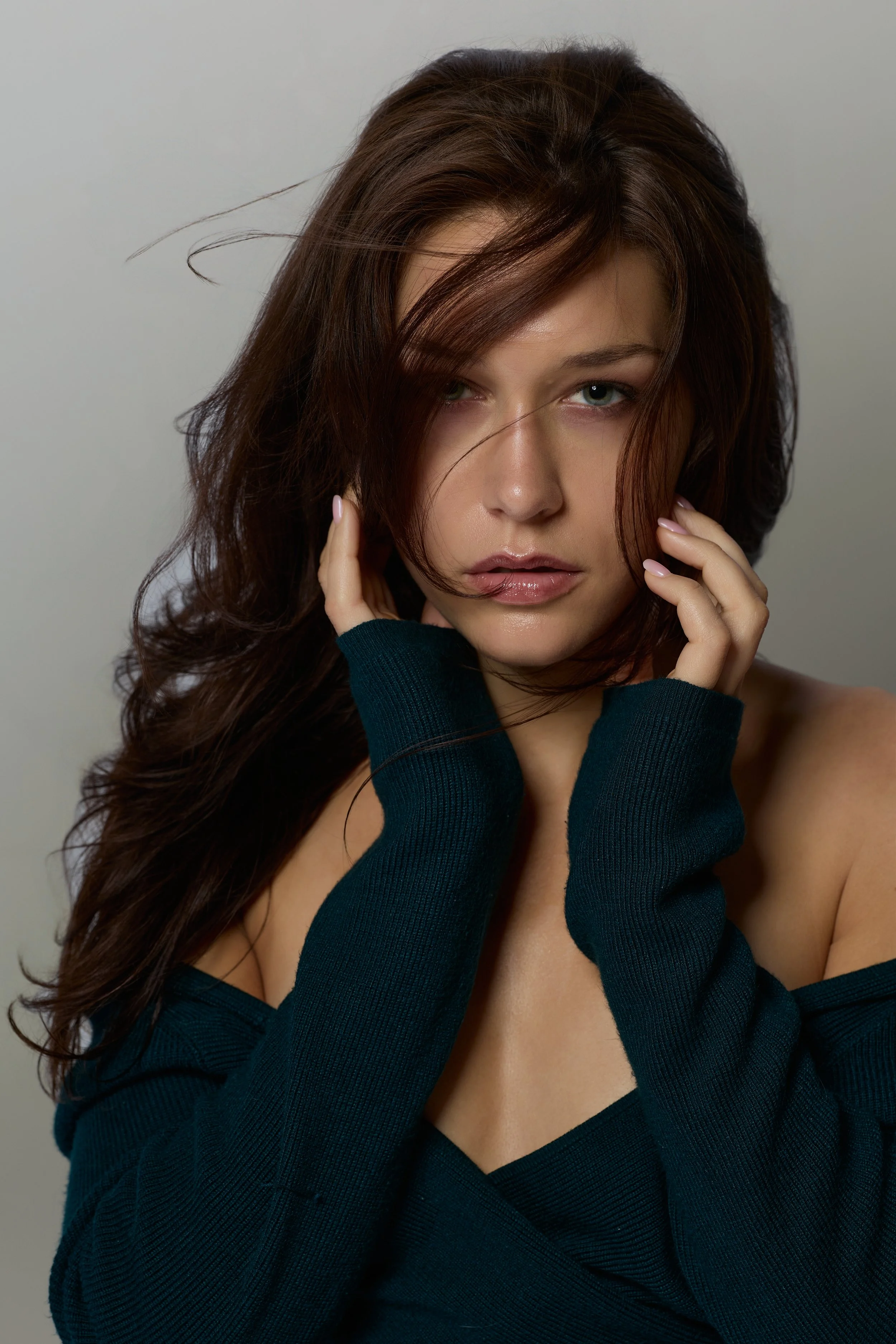 Close-up of a young woman with long wavy brown hair, touching her face with both hands, wearing a dark teal off-the-shoulder top, against a plain light grey background.