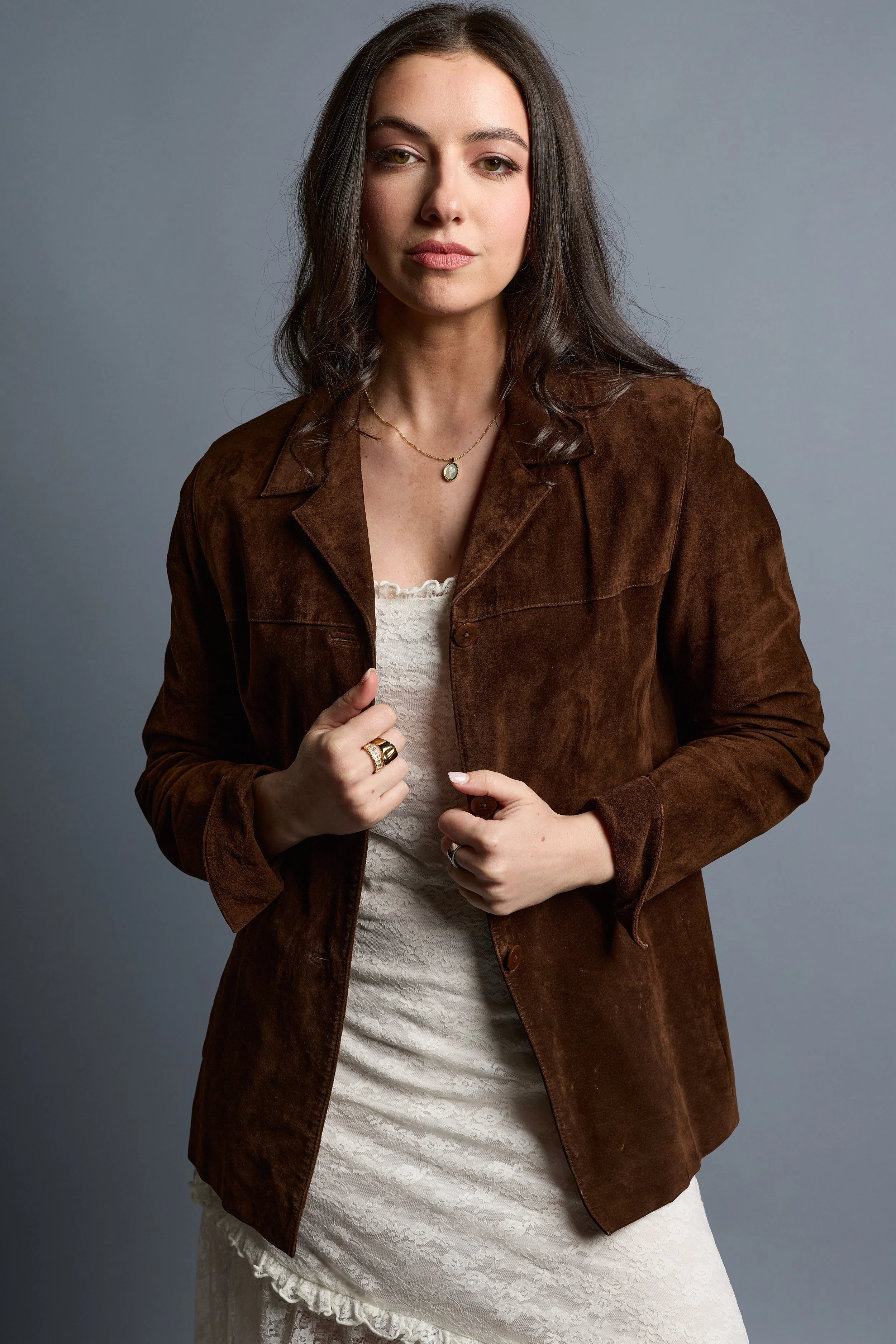 A young woman with shoulder-length dark brown hair, wearing a brown jacket over a white lace dress, accessorized with a small pendant necklace and rings on her fingers, standing against a plain gray background.