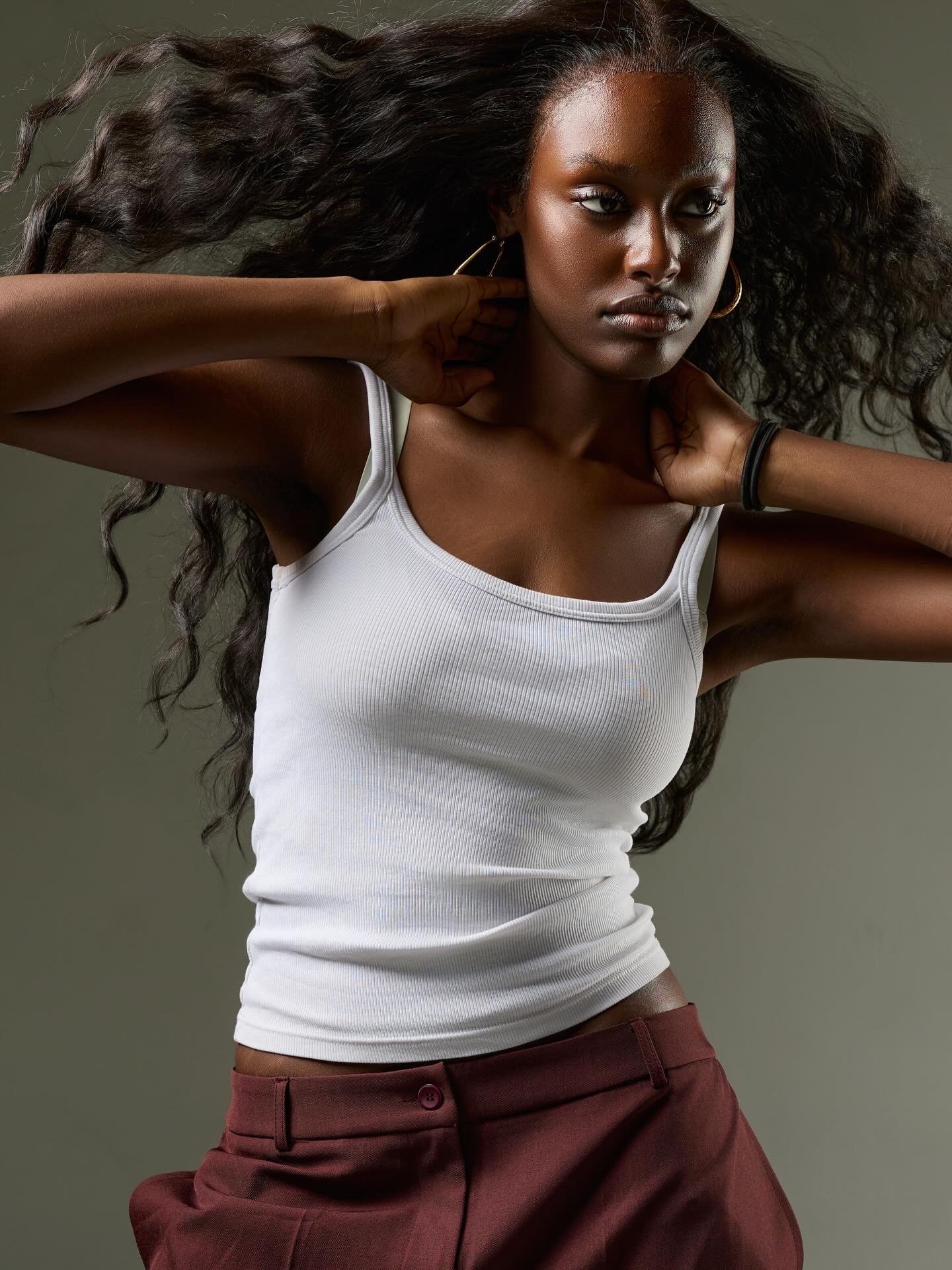 A woman with dark skin and long, curly hair is wearing a white tank top and maroon pants. She has one hand behind her neck and the other near her ear, wearing large hoop earrings and a black bracelet. The background is plain and neutral.