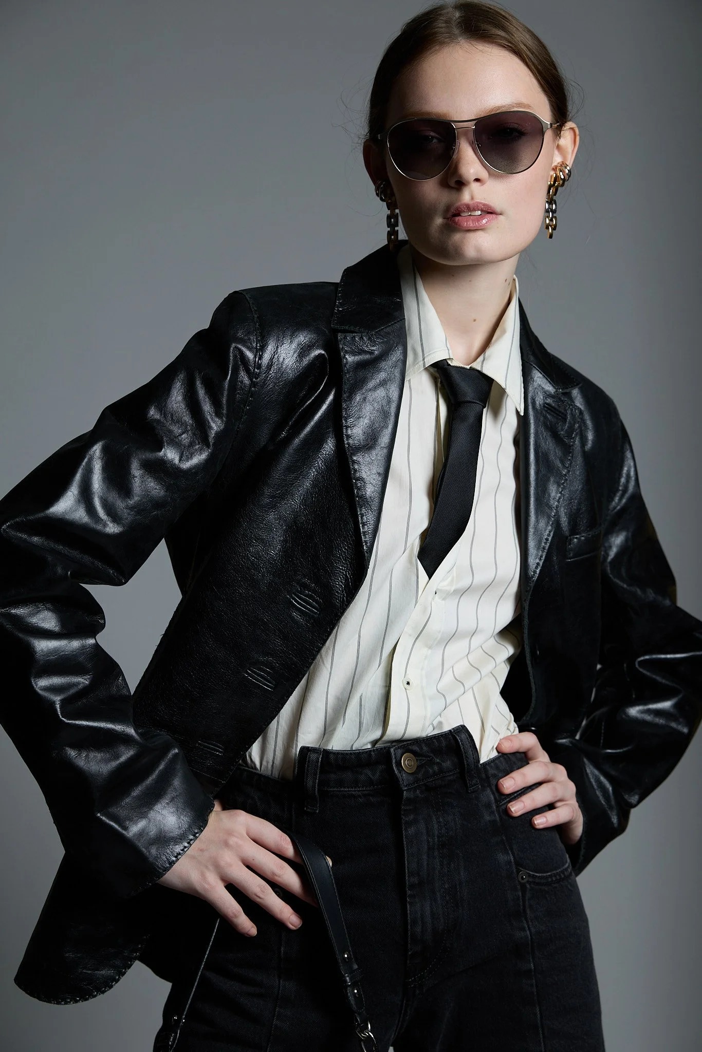 Woman wearing sunglasses, a black leather jacket, a striped button-up shirt, and black jeans, standing with hands on hips against a neutral background.