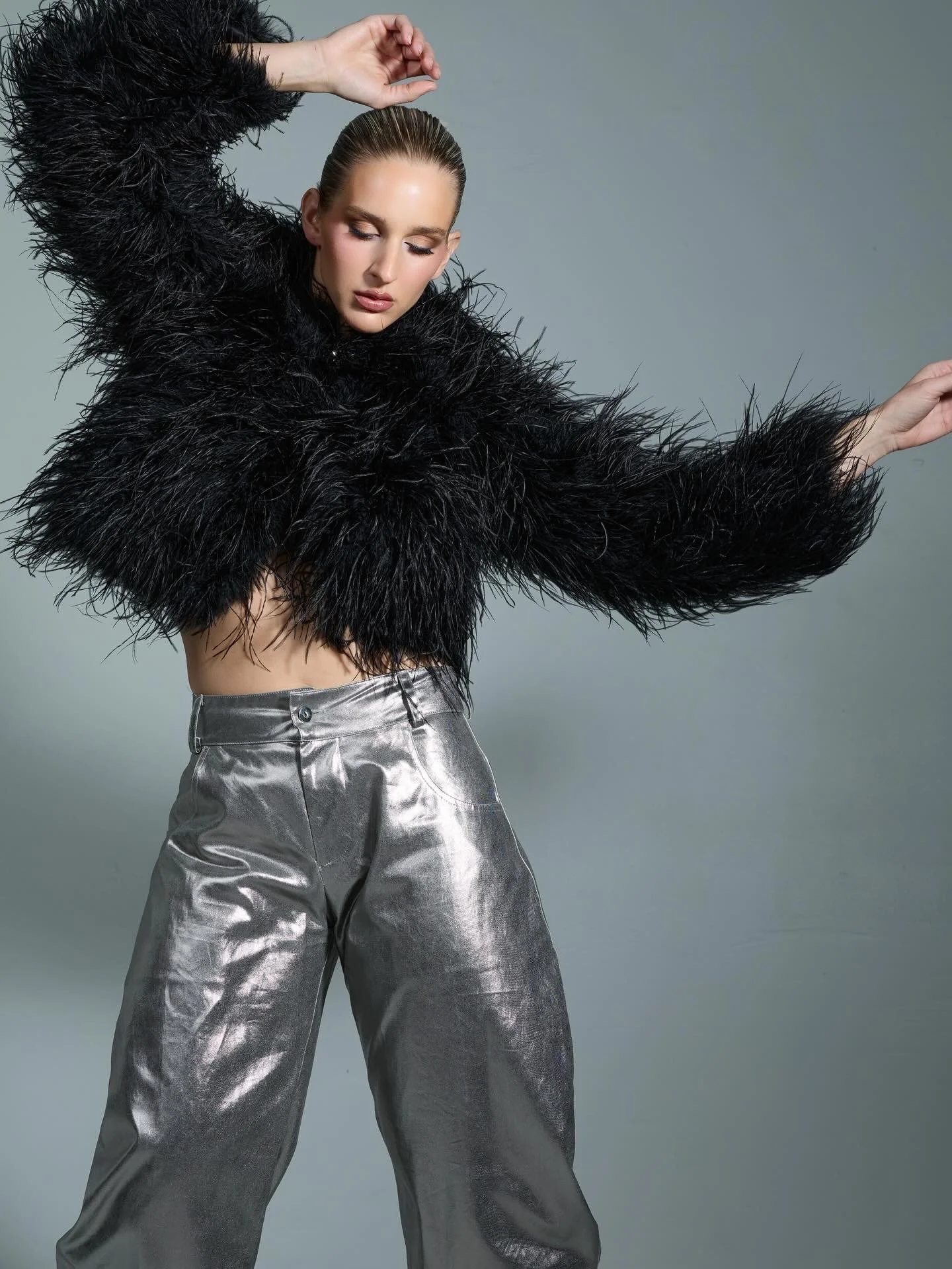 A woman with slicked-back hair is wearing a black furry jacket and shiny silver pants, posing with her eyes closed against a plain gray background.
