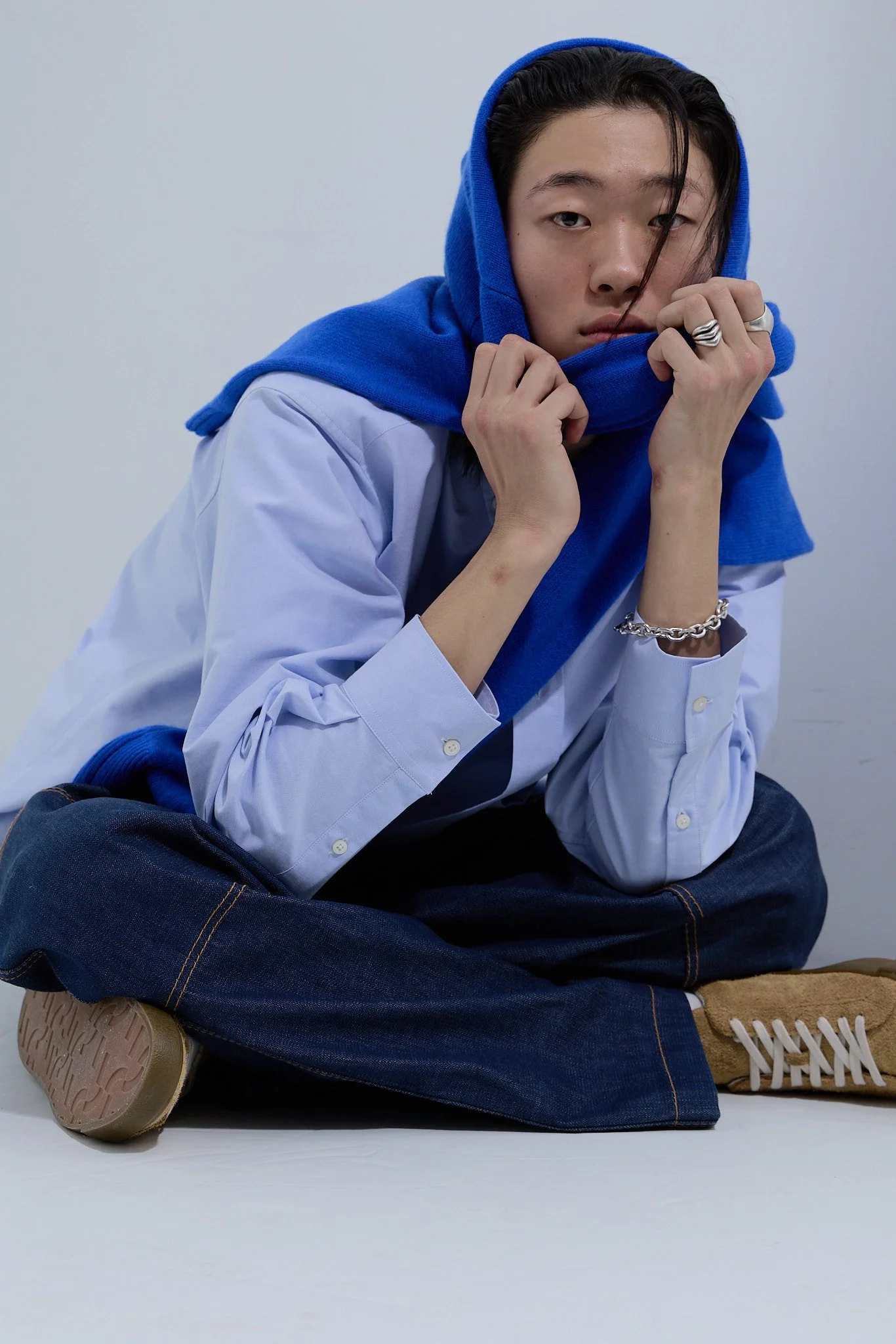 A young man sitting cross-legged on the floor, wearing a white shirt with rolled-up sleeves, a blue hoodie draped over his head, and dark jeans. He has rings and a bracelet, and is holding the hoodie with both hands while looking into the camera with