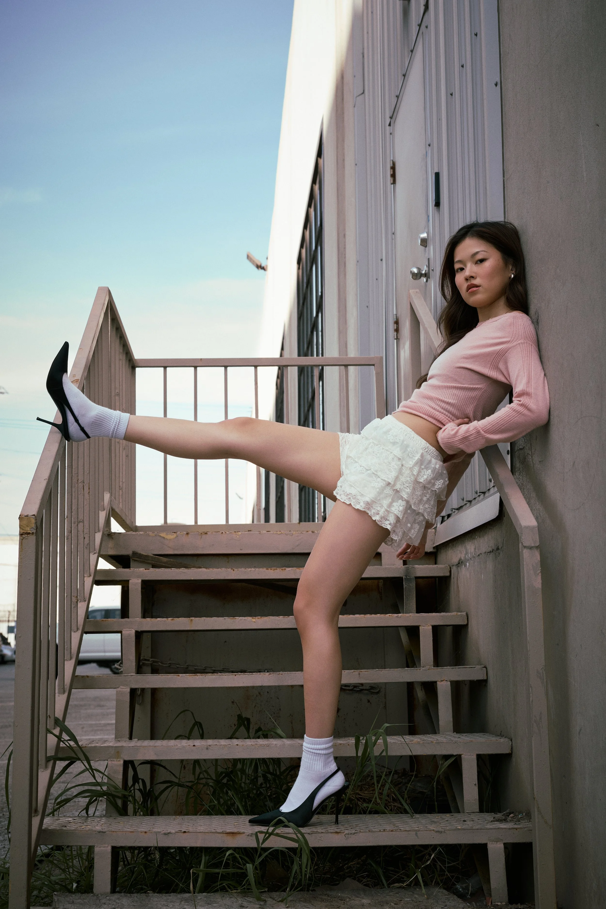 Young woman with long dark hair sitting on outdoor stairs, wearing a pink long-sleeve top, white lace shorts, white socks, and black high heels, leaning against a wall with a serious expression.