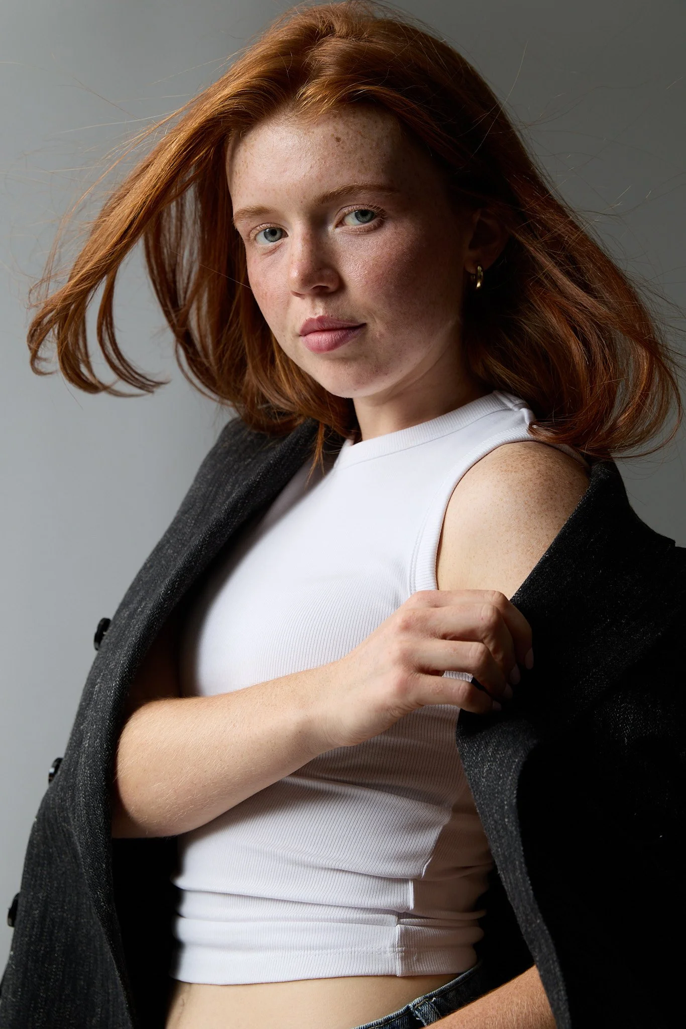 Young woman with red hair, wearing a white sleeveless top and a black blazer, holding her blazer open to reveal her shoulder, against a plain background.