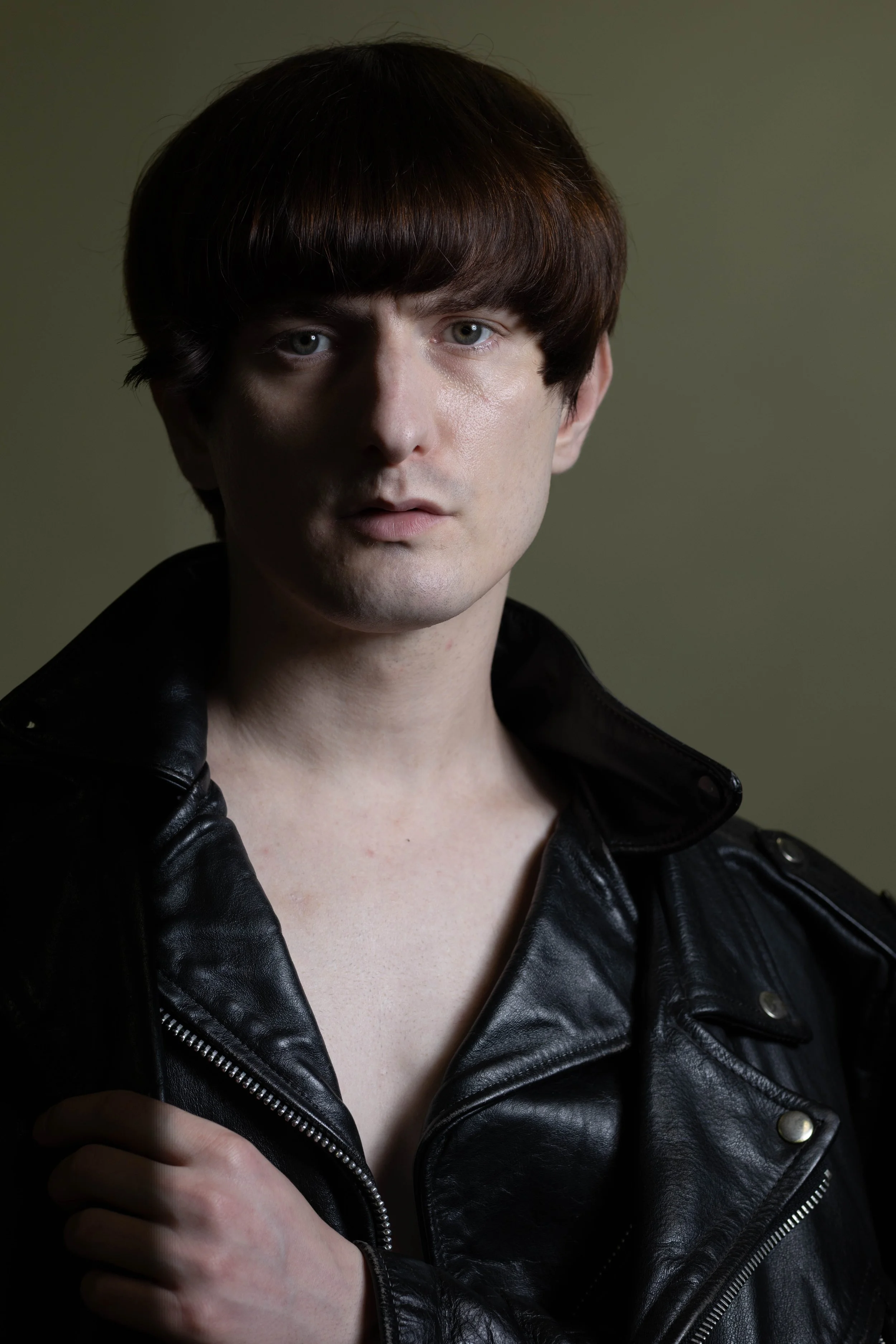 A young man with straight brown hair and fair skin, wearing a black leather jacket, poses against a plain gray background.