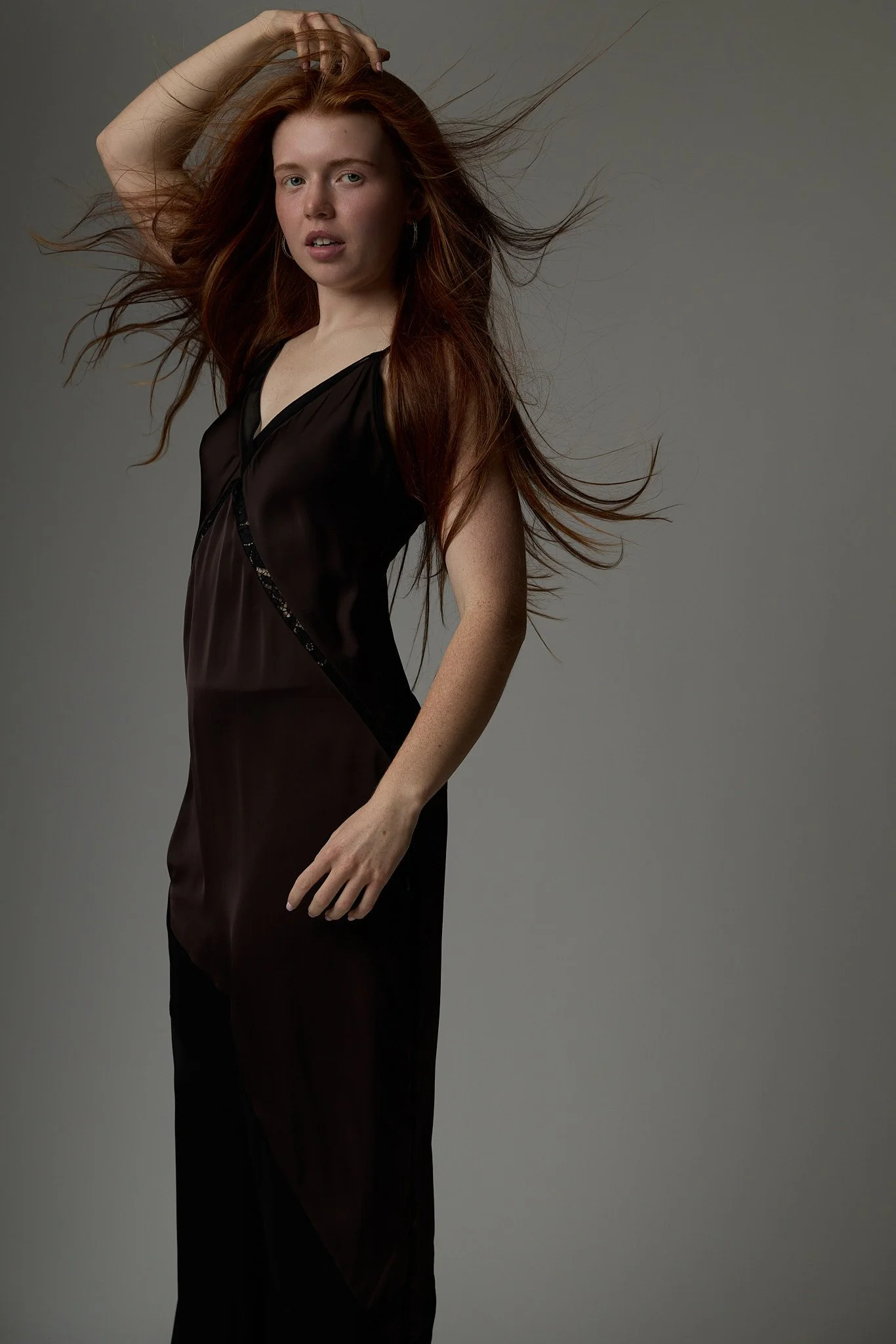 A young woman with long red hair, wearing a black satin slip dress, posing against a gray background with her hand running through her hair.