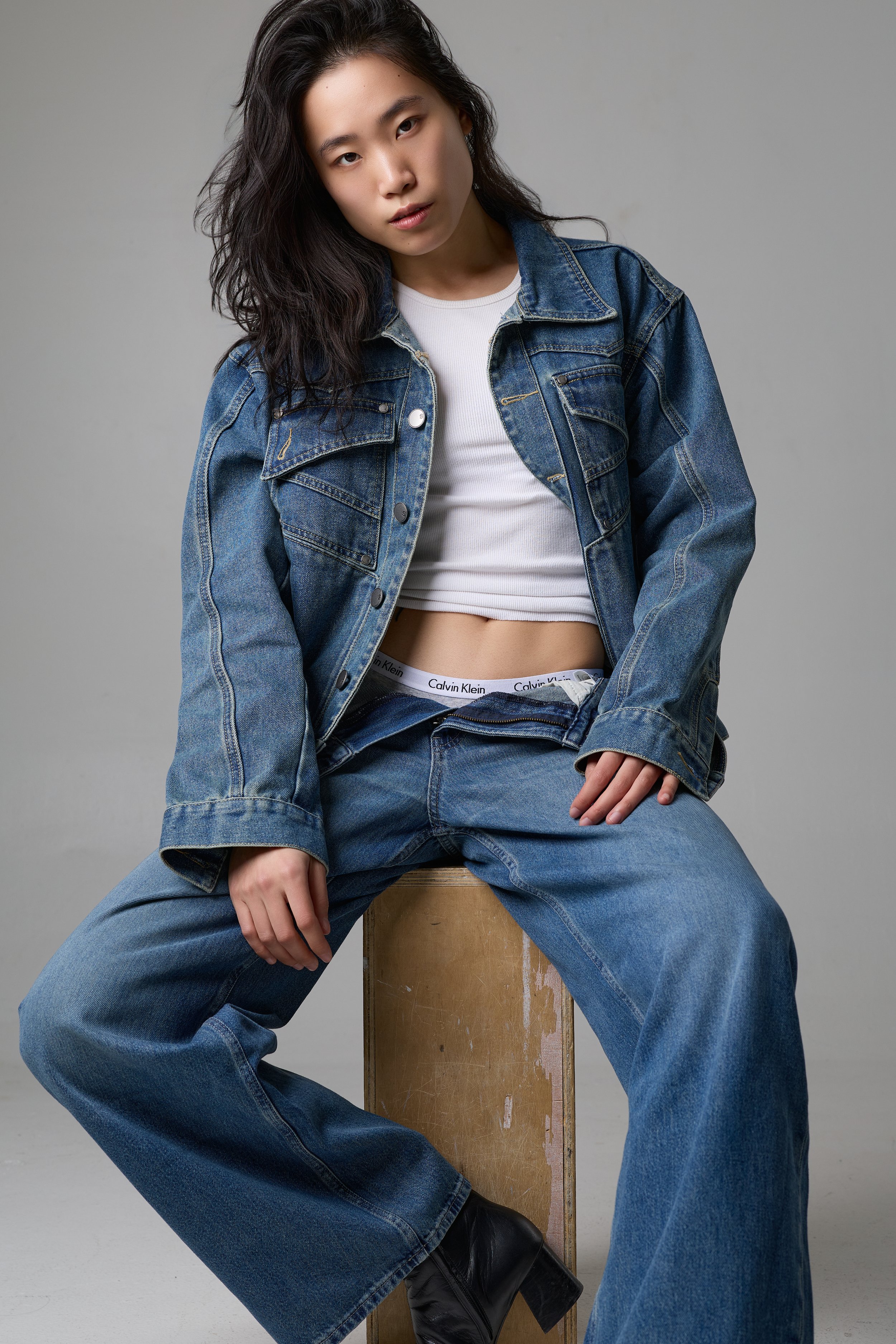 A young woman with long, dark, wavy hair wearing a denim jacket over a white crop top, blue jeans, and black boots, sitting on a wooden block against a plain gray background.
