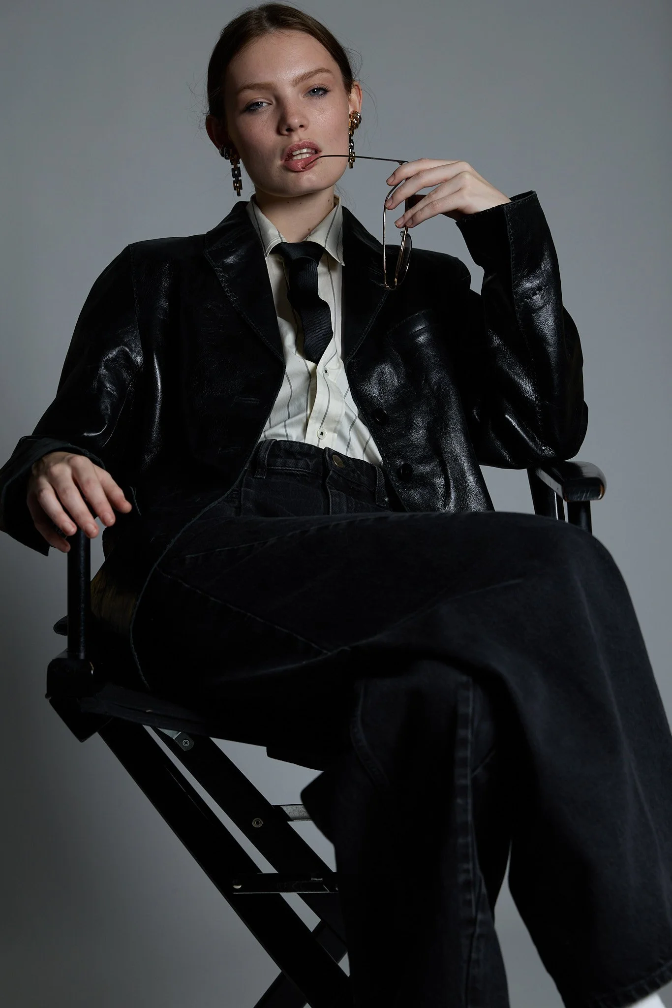 Young woman with auburn hair seated in a black folding chair against a plain gray background, wearing a black leather jacket, white shirt, black tie, and black pants, holding glasses and biting the earpiece.