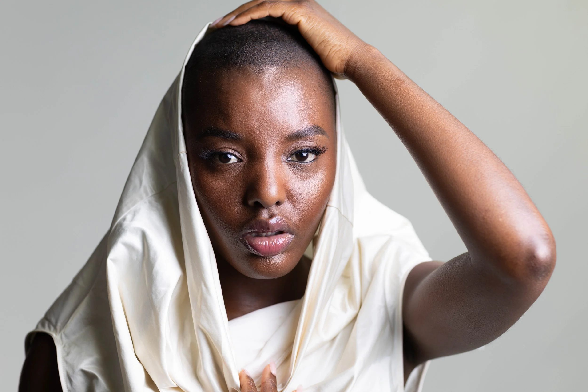A woman with dark skin, short hair, wearing a white sleeveless top with a cowl neckline, holding her head with one hand, looking directly at the camera with a neutral expression.