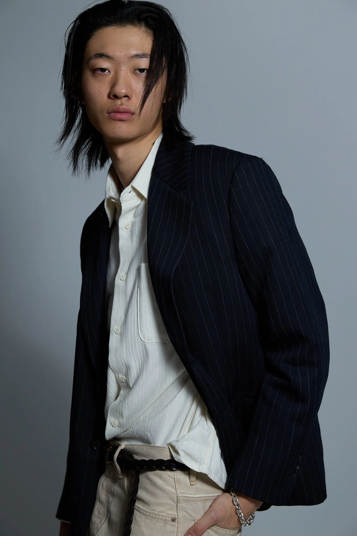 A young man with long black hair, wearing a dark pinstripe blazer over a white shirt, beige pants, and a silver bracelet.