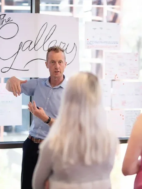 Man giving a presentation to two women in an office with a whiteboard and notes.