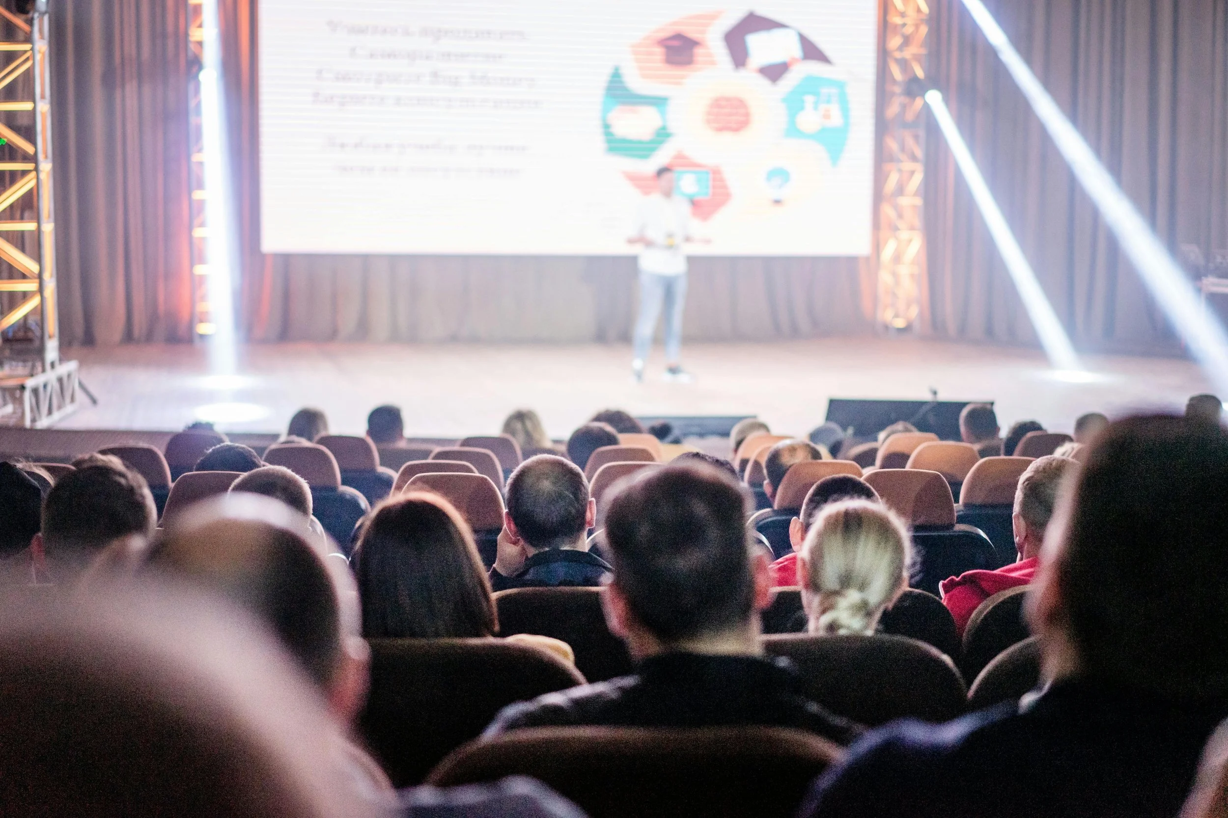 A speaker presenting on stage at a conference with an audience seated in rows. The presentation slide behind shows colorful icons and text.