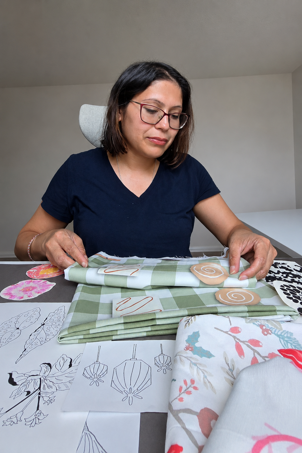 Pratyasha Sharma, founder of Align One Surface Pattern Studio, sitting at a table folding fabric, surrounded by floral and leaf-patterned cloth and sketches of holiday lights and pinecones.