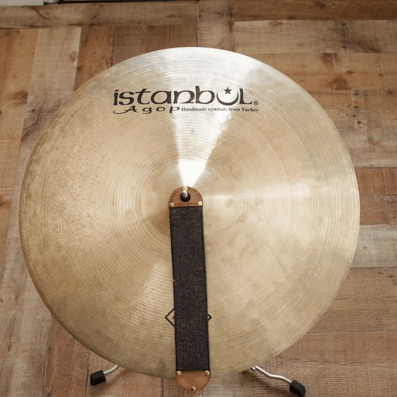 A large golden cymbal with the brand name 'Istanbul Agop' printed on it, placed on a wooden floor, with a felt strip and a metal post at its center.